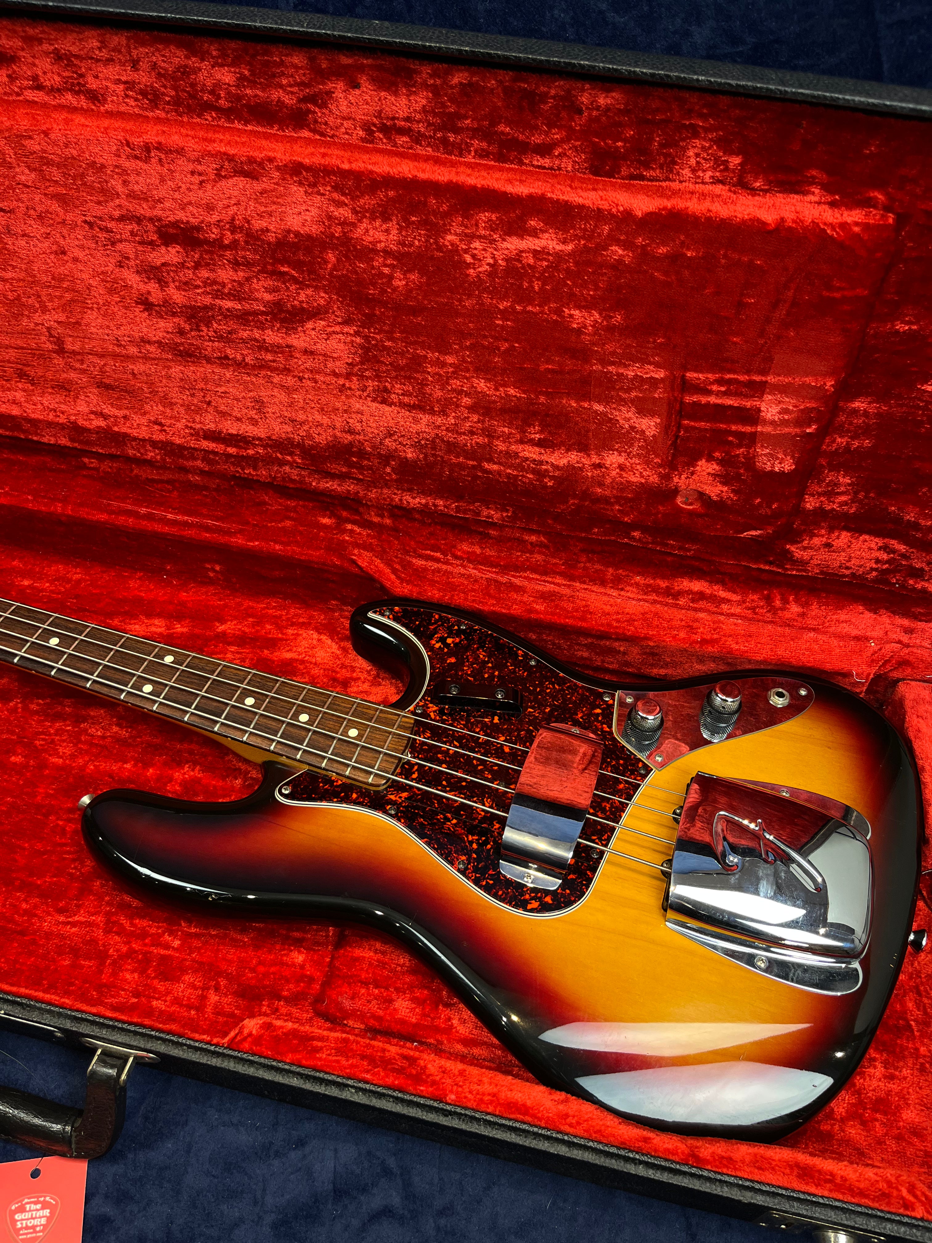 Sunburst-finish bass guitar with tortoiseshell pickguard and chrome covers, lying in a plush red velvet-lined hard case.