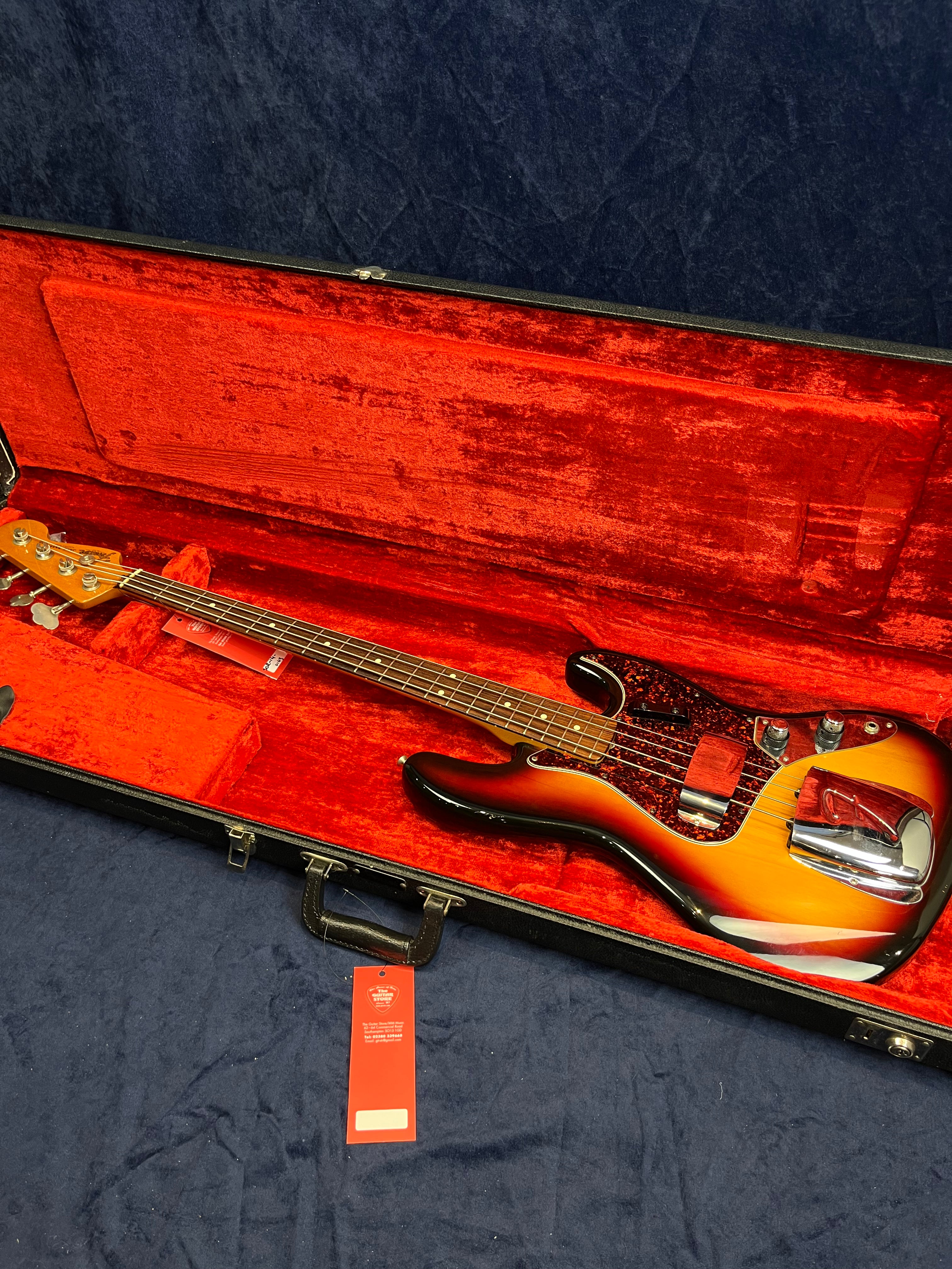 Sunburst finish bass guitar with chrome covers, tortoiseshell pickguard, and four strings, resting in a red velvet-lined hard case.