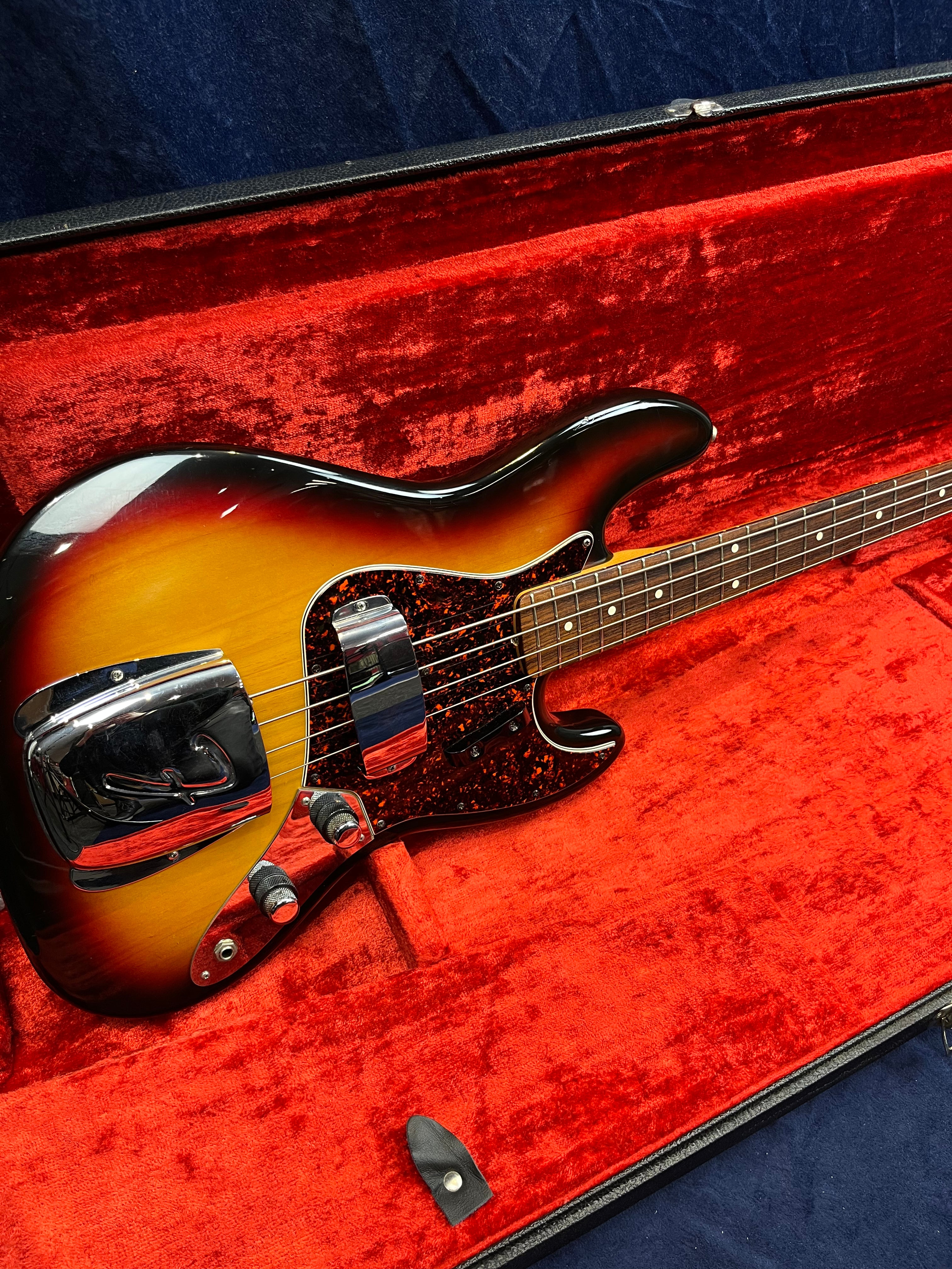 Sunburst finish bass guitar with chrome pickup covers and tortoiseshell pickguard, resting in a plush red-lined hard case.