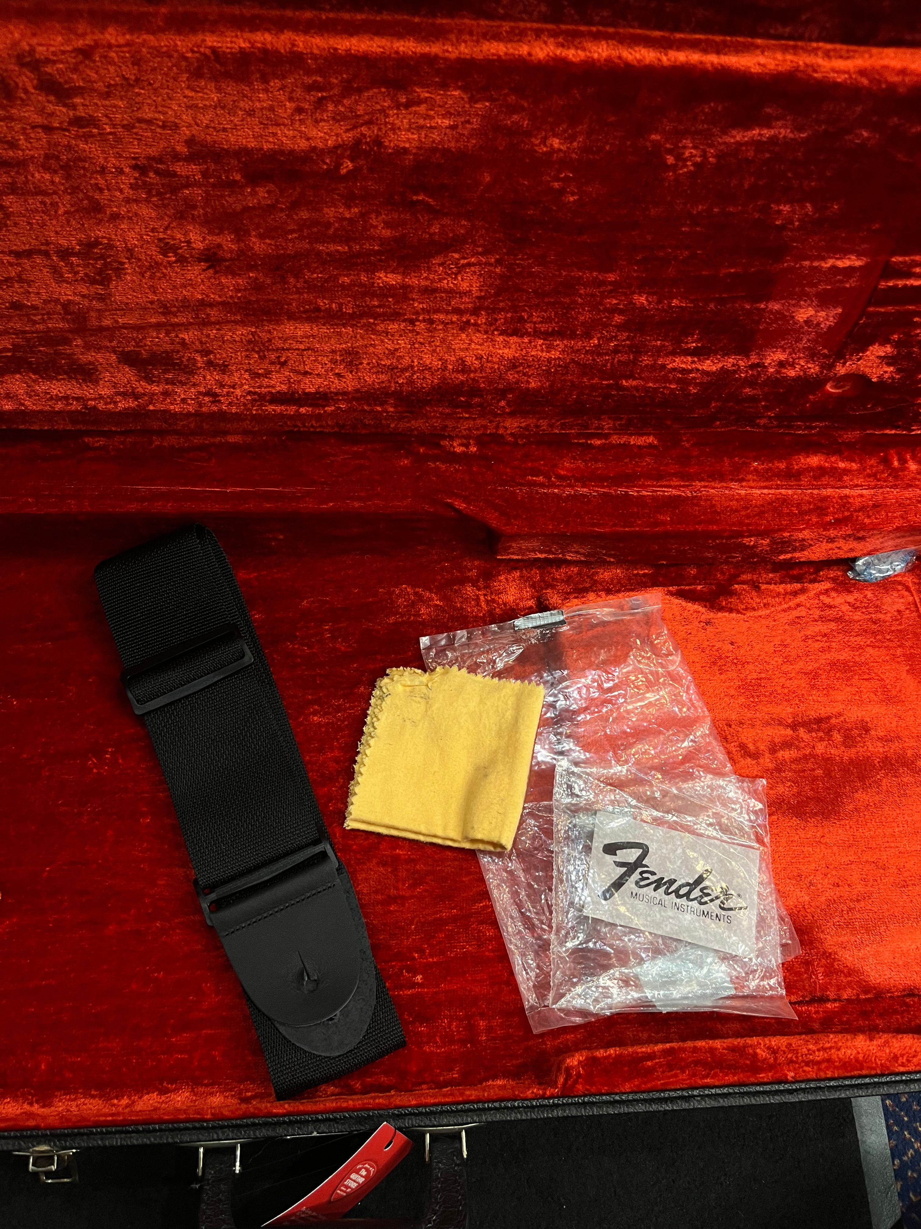 Black guitar strap, yellow cleaning cloth, and a clear Fender-branded bag inside a red velvet-lined bass guitar case.