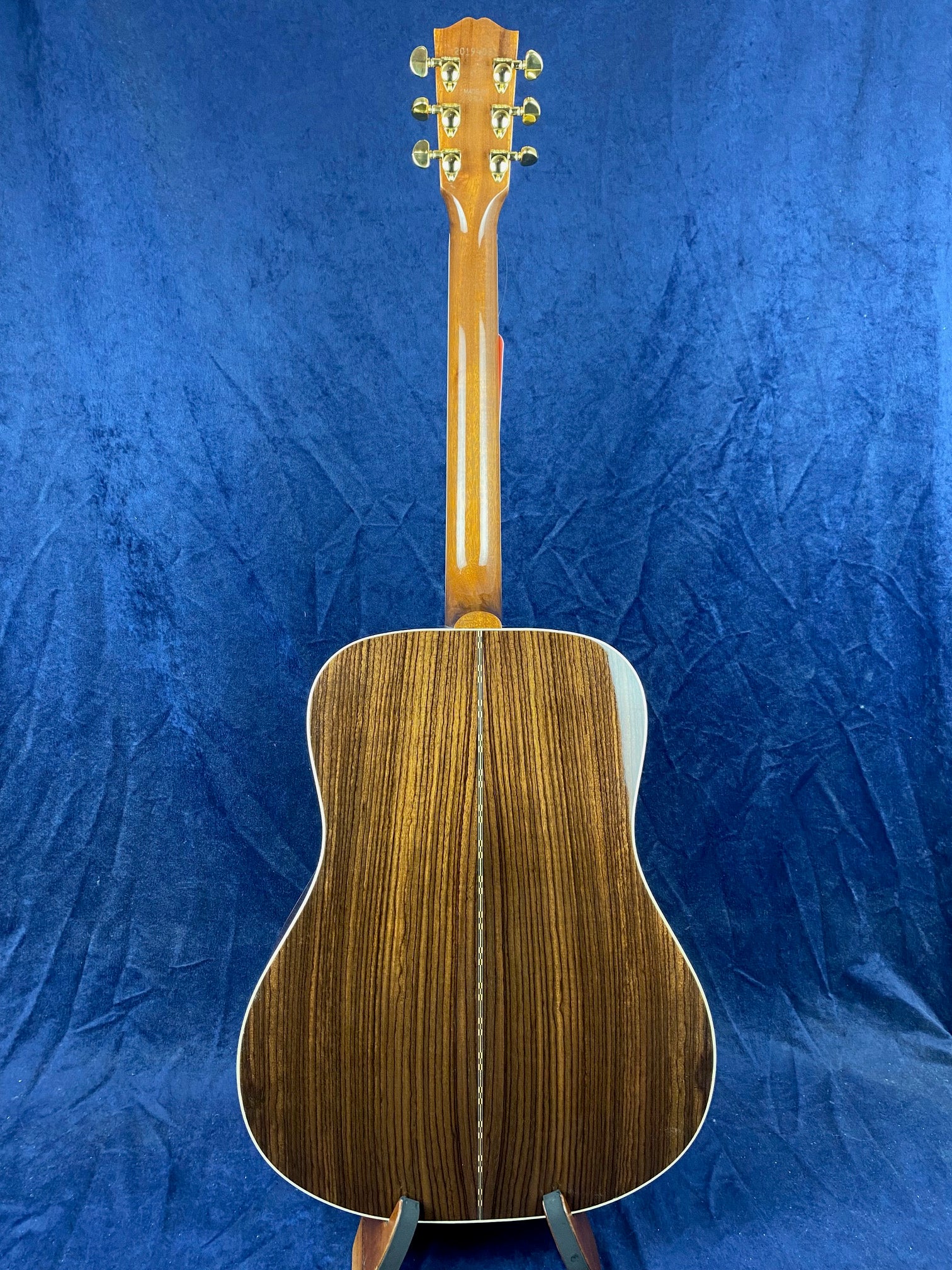 The back of an acoustic guitar with a glossy, dark wood finish featuring vertical grain patterns and gold tuning pegs, set against a blue background.