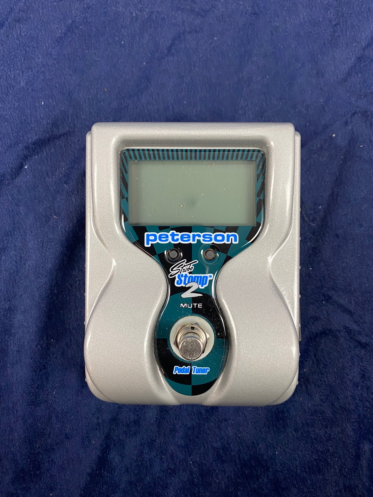 Silver Peterson Stomp 2 pedal tuner with a footswitch and large screen, labeled “Mute” and “Pedal Tuner,” resting on a dark blue fabric.
