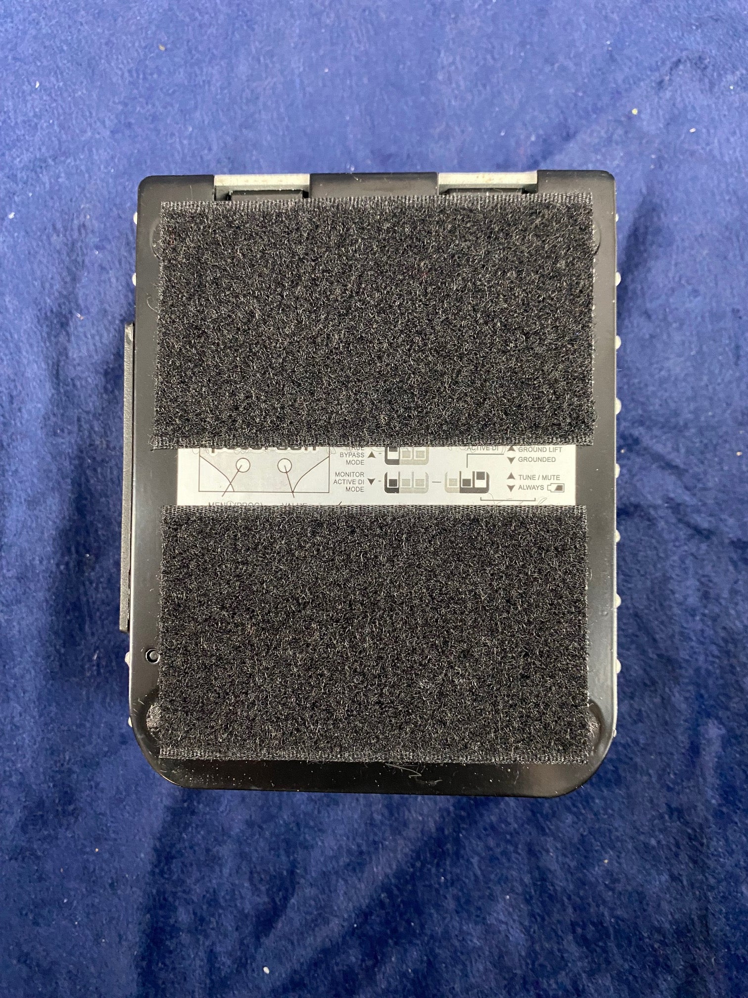Rectangular guitar effects pedal, bottom view, with two large black Velcro strips covering most of the surface against a blue background.