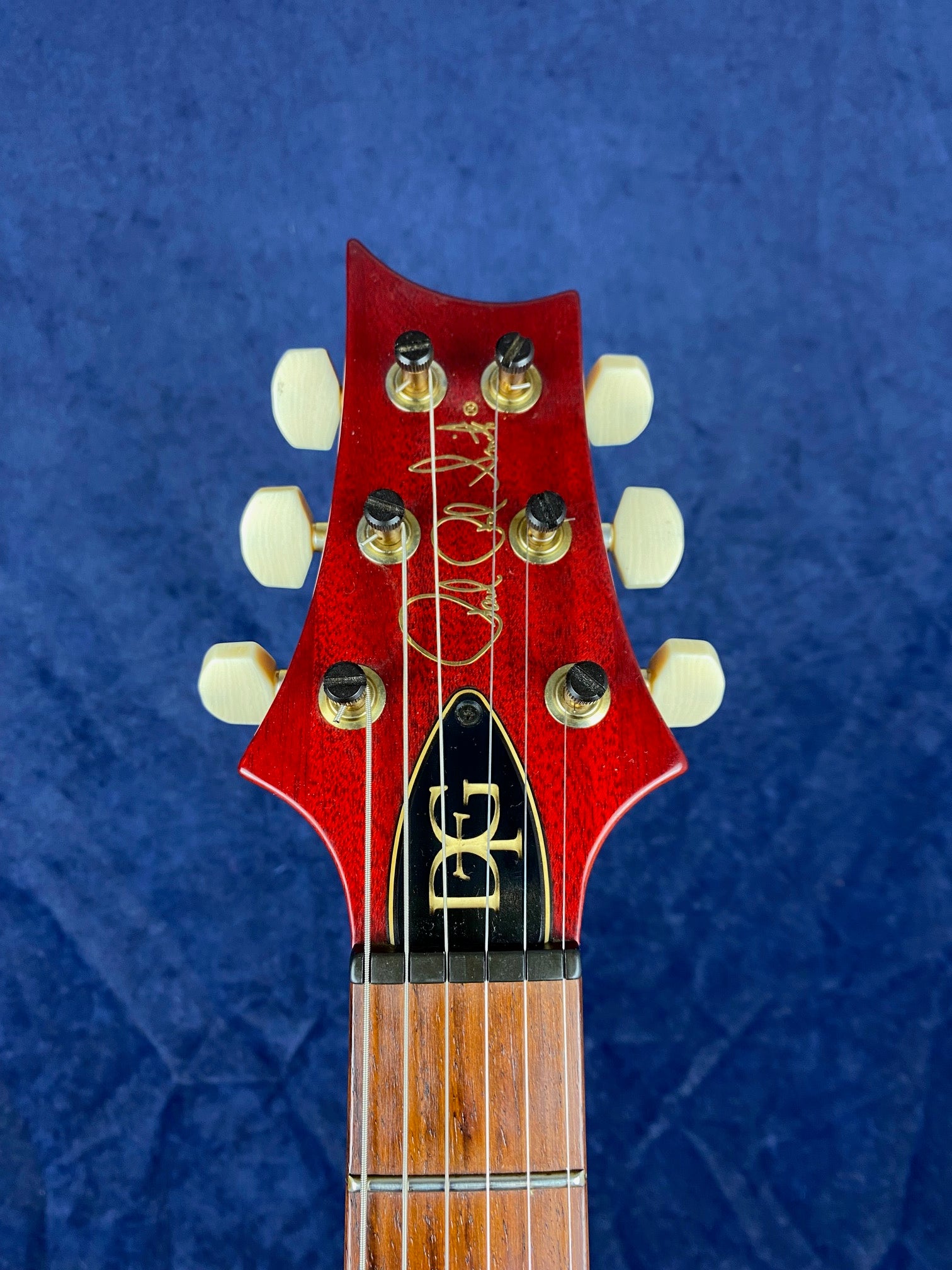 Electric guitar headstock in glossy red with six cream tuning pegs, gold signature, and an inlaid gold “EG” logo on a black truss rod cover.