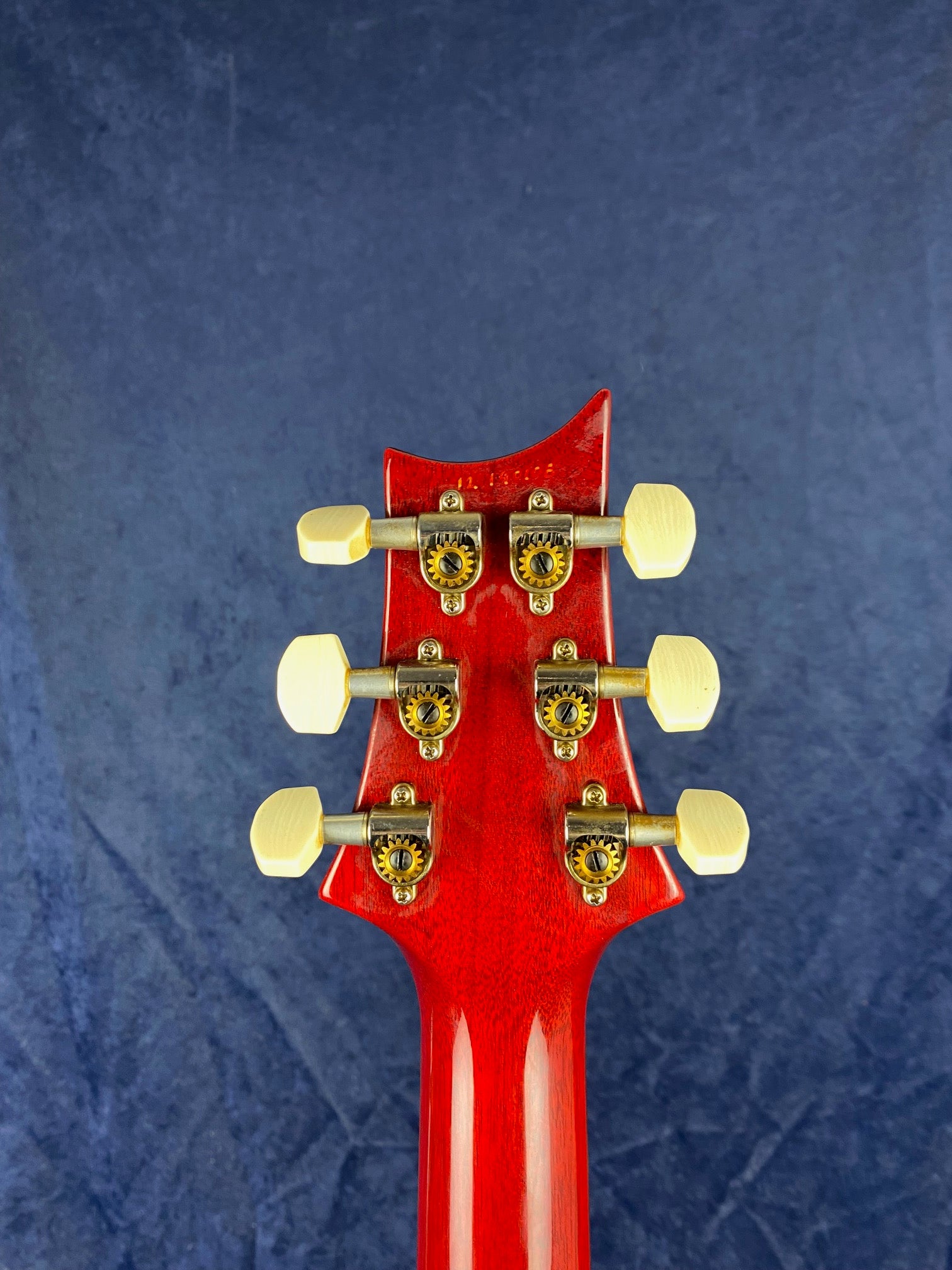 Back of a red electric guitar headstock showing six gold tuning pegs with ivory-colored knobs against a dark blue background.