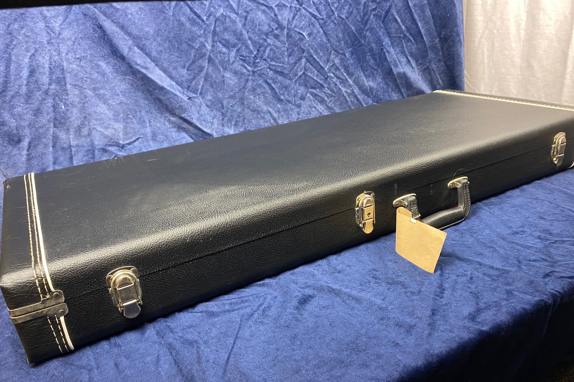 Rectangular black electric guitar hard case with metal latches, a handle, and a tan tag, resting on a blue fabric surface.