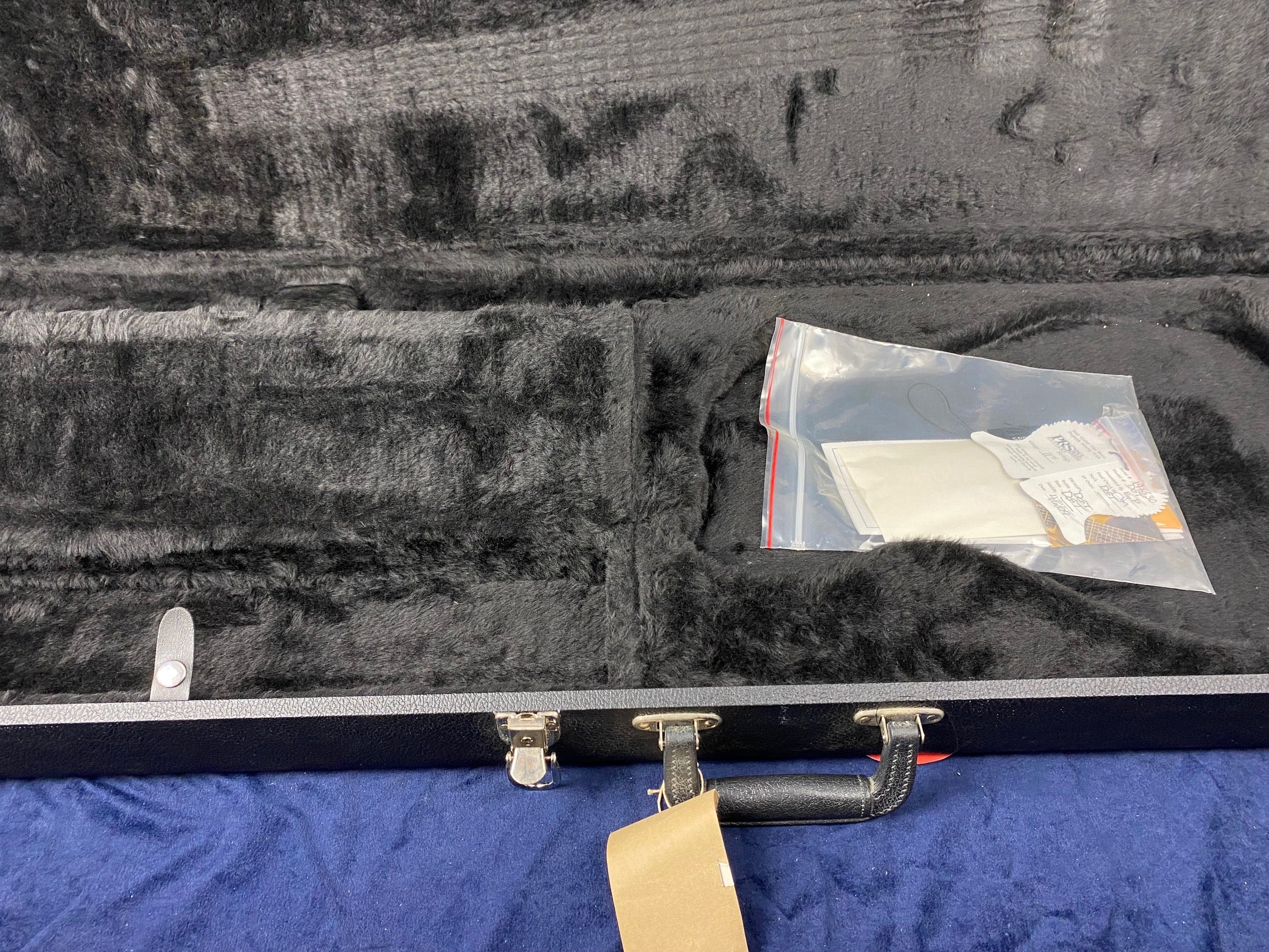 Empty black plush-lined electric guitar case with a plastic bag containing papers and a cloth inside the accessory compartment.