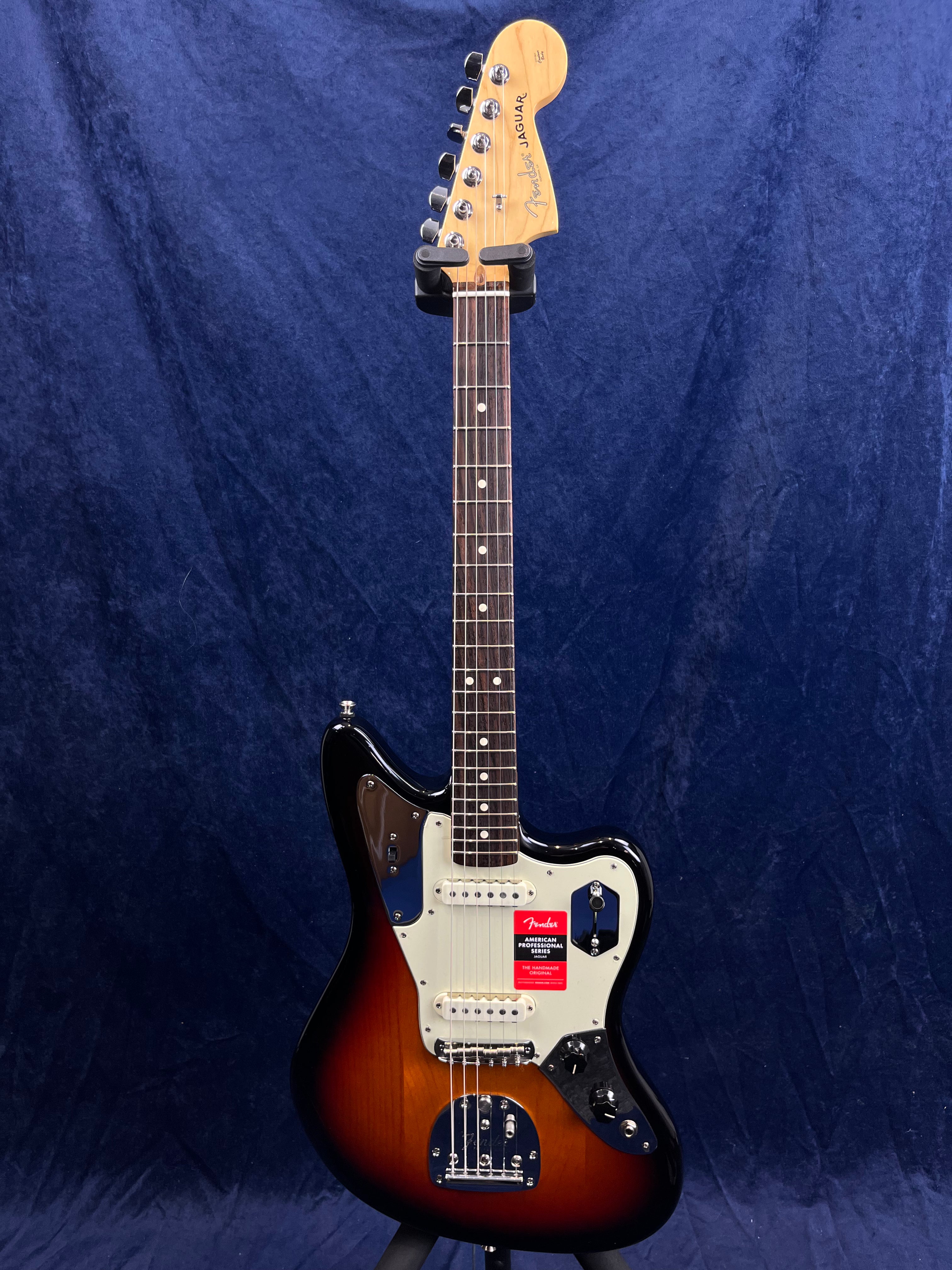 Sunburst Fender Jaguar electric guitar with a white pickguard, two pickups, multiple control knobs, and a rosewood fretboard against a dark blue background.
