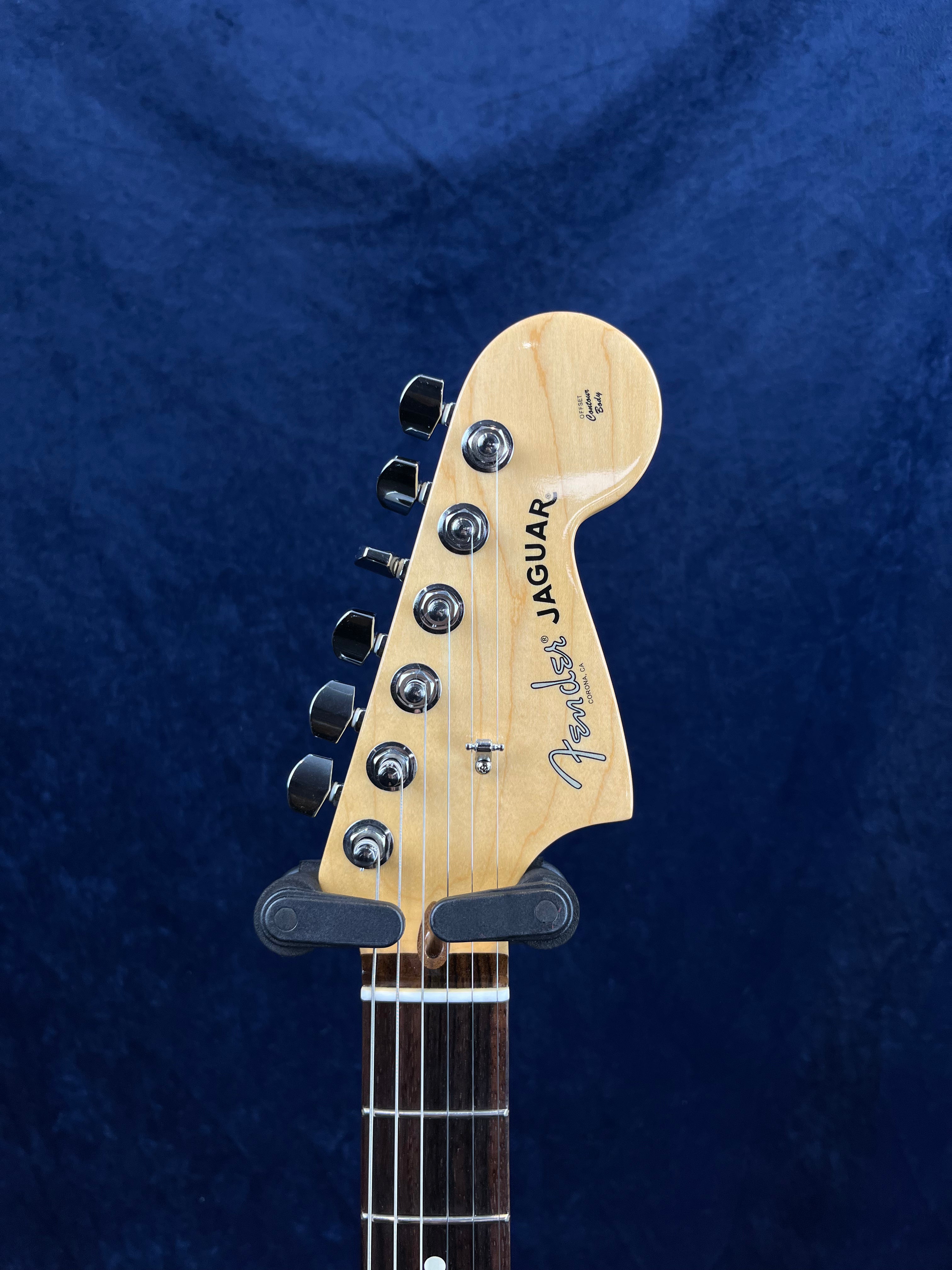 Light wood Fender Jaguar electric guitar headstock with six tuning pegs and "Jaguar" and "Fender" logos, set against a dark blue background.