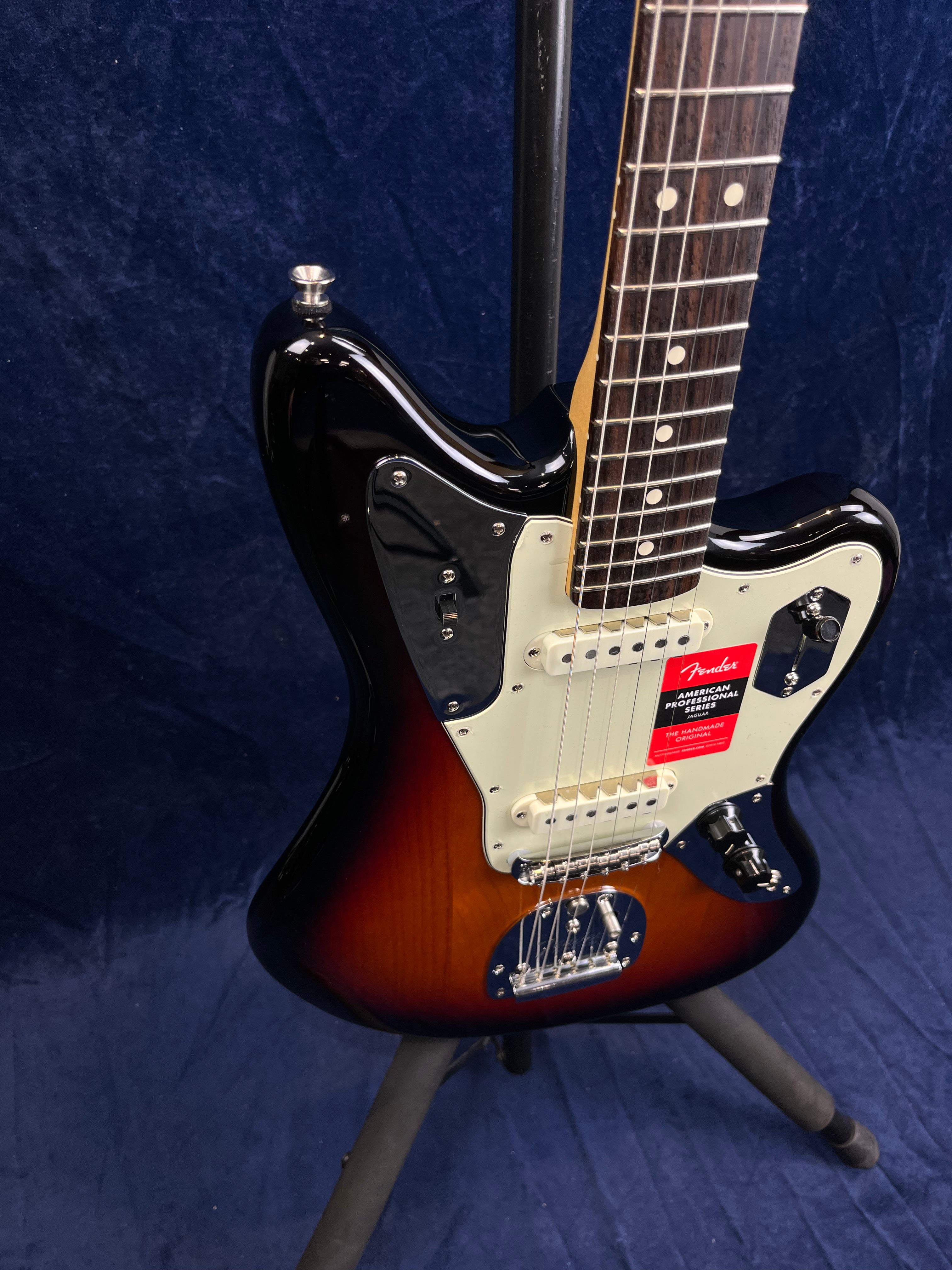 Sunburst Fender electric guitar on a stand, white pickguard, dark fretboard with dot inlays, red label near pickups, blue background.