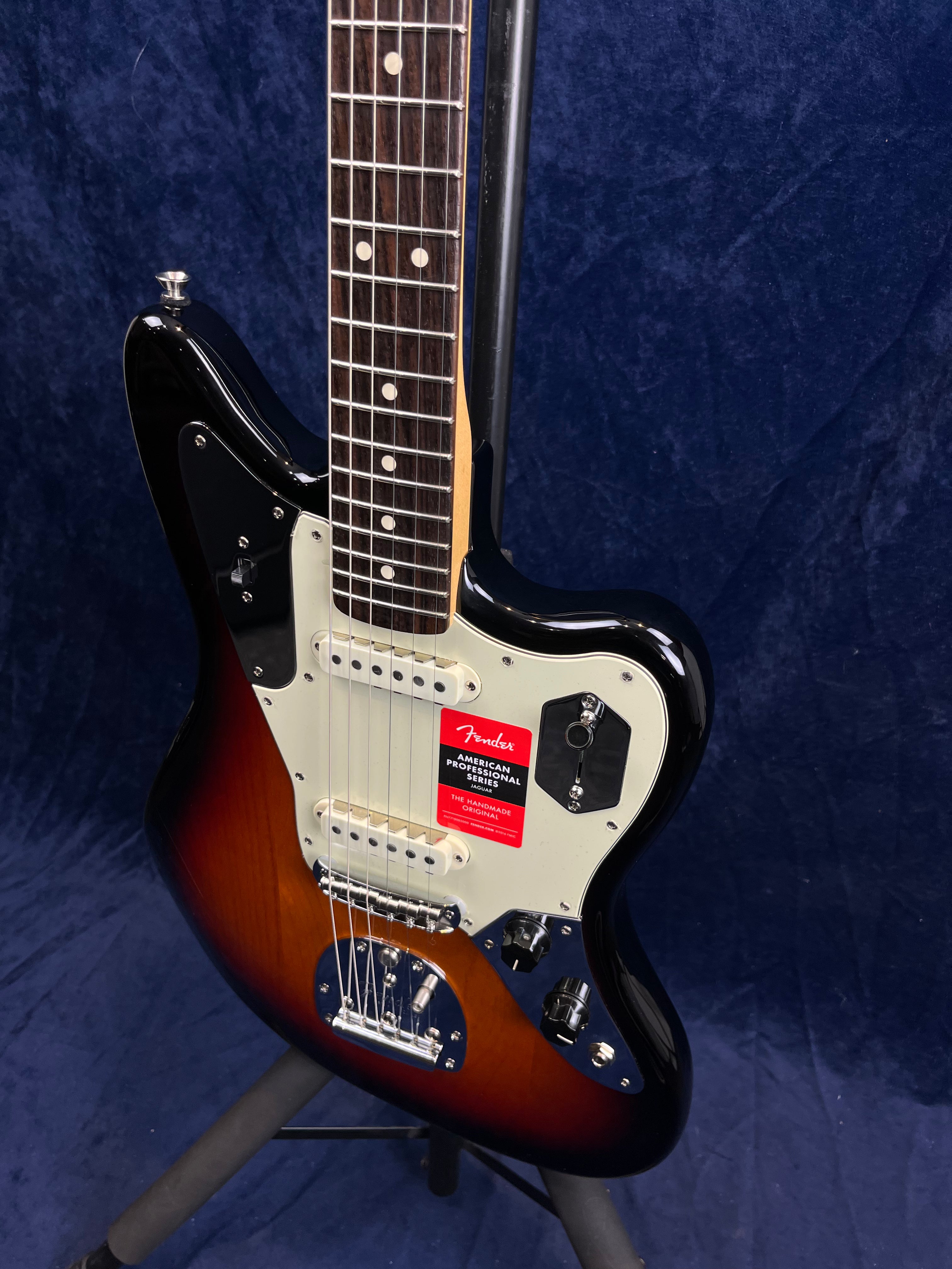 Sunburst Fender electric guitar with a white pickguard, two pickups, dark fretboard, and control knobs, displayed on a stand in front of dark fabric.