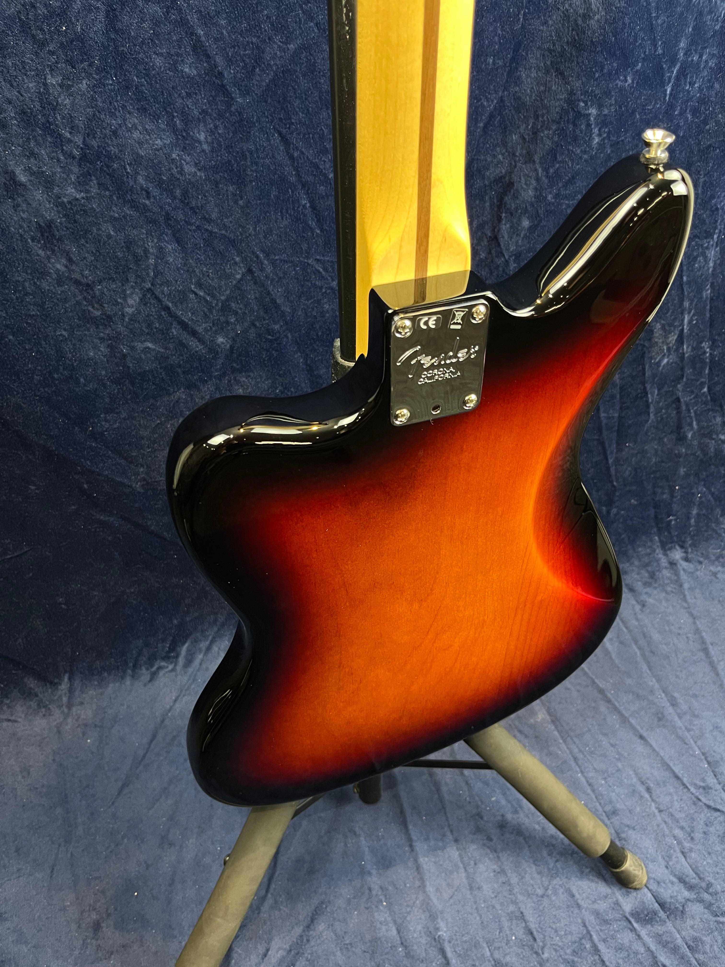 Sunburst-finish electric guitar showing its contoured back, bolt-on maple neck, and metal neck plate with the Fender logo, resting on a stand.