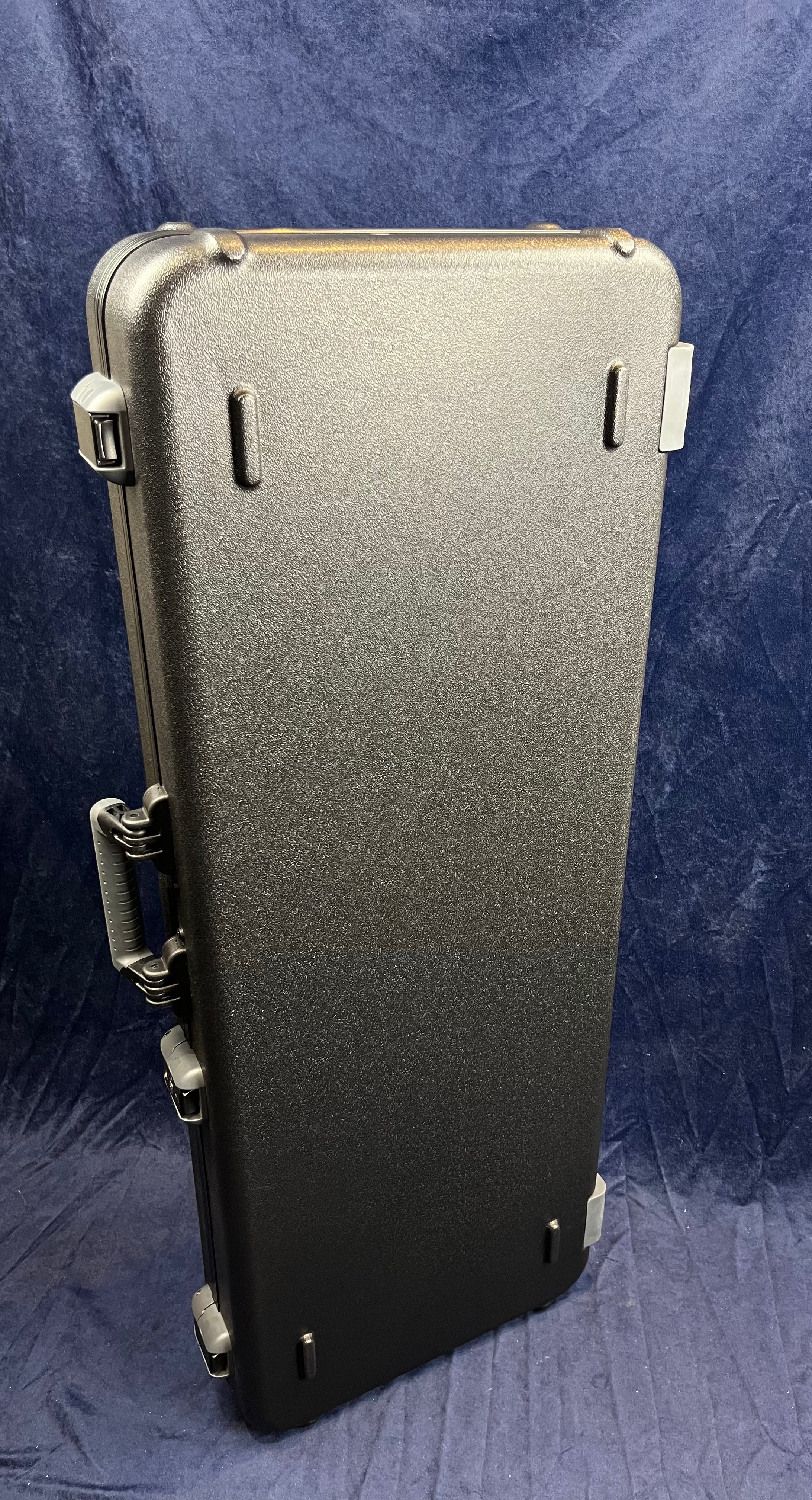 A hard-shell black electric guitar case stands upright, with four latches, molded handle, and slightly textured surface against a dark fabric background.