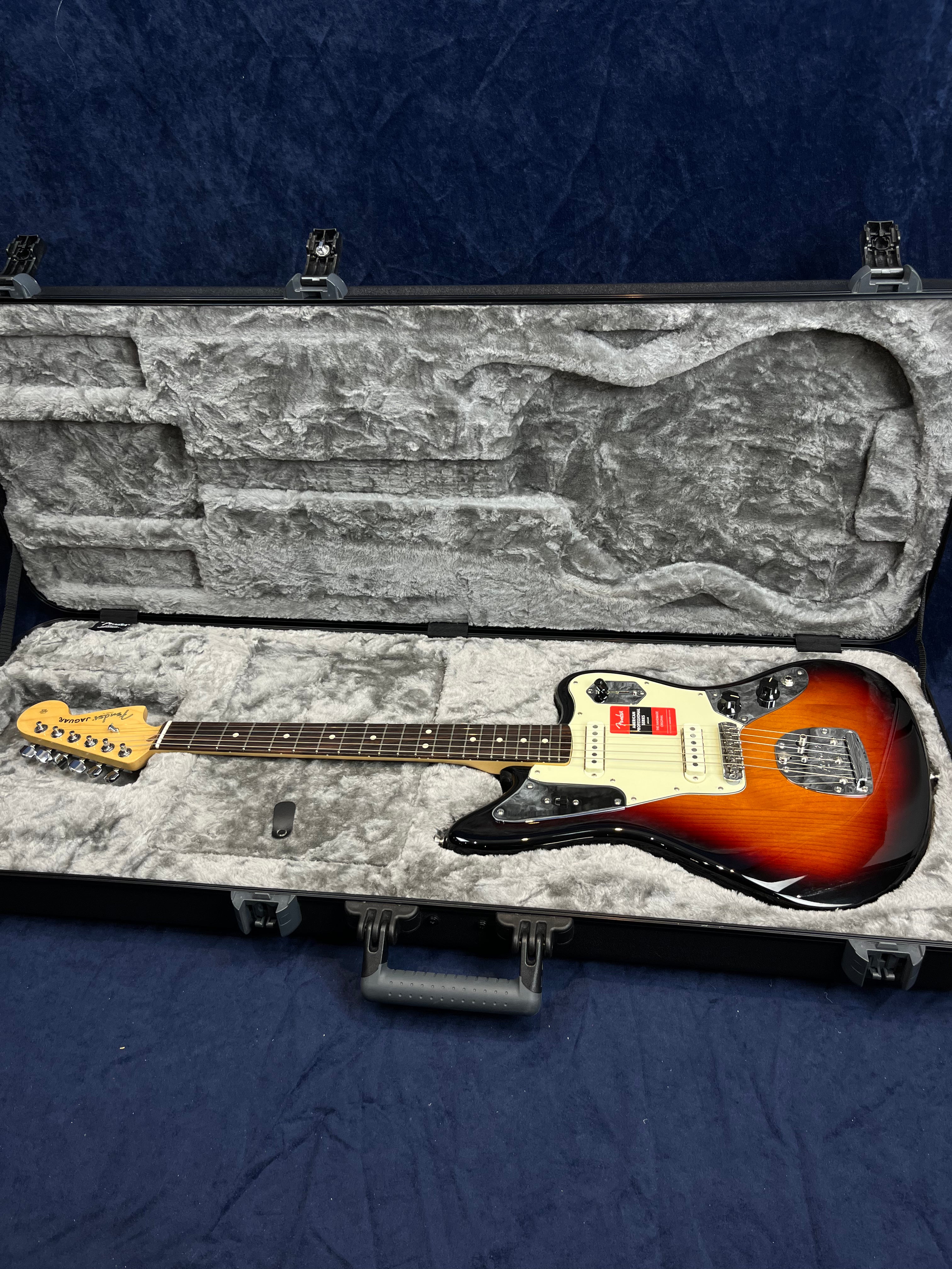 Sunburst Fender Jaguar electric guitar with white pickguard in a hard gray plush-lined case, all resting on a dark blue surface.
