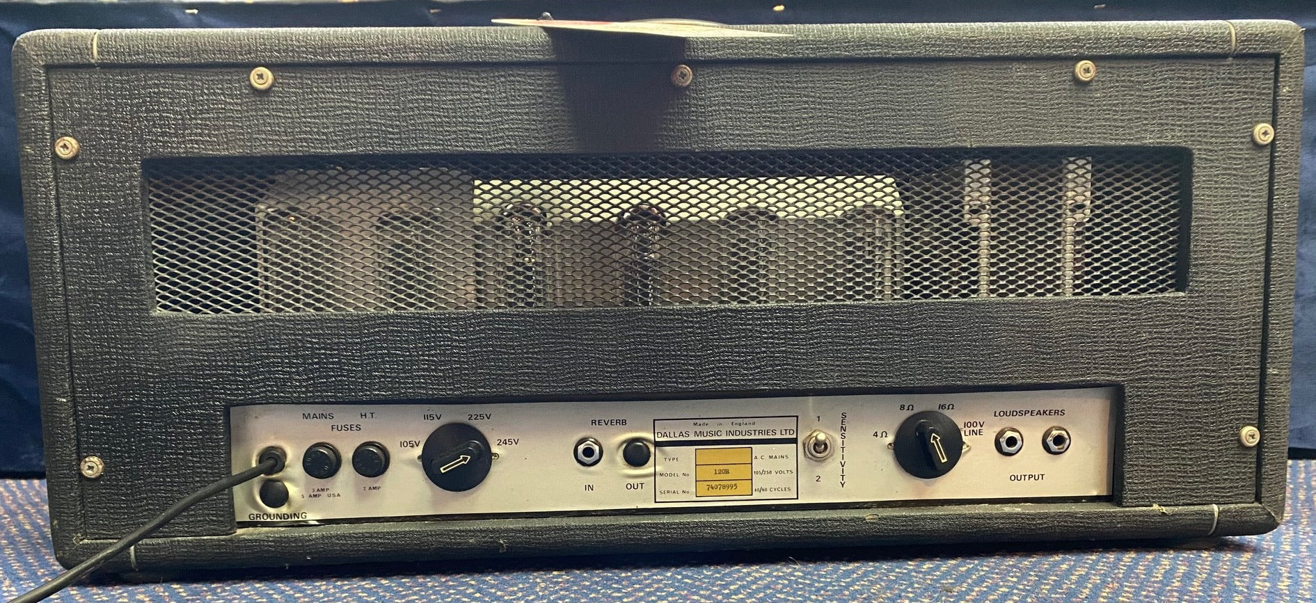 Black guitar amplifier head with visible vacuum tubes behind a metal grille, multiple control knobs, fuse ports, input/output jacks, and voltage selector.