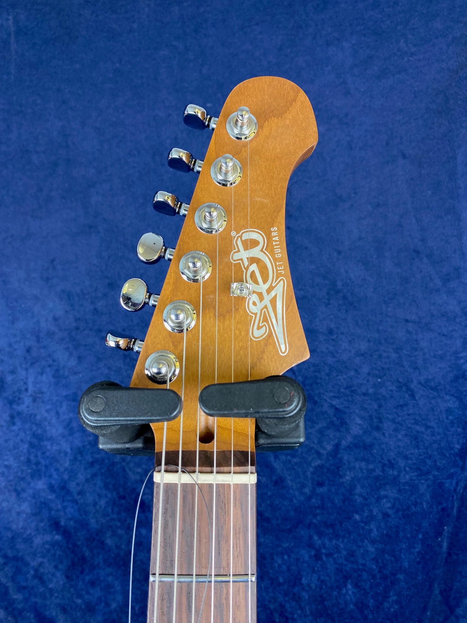 Electric guitar headstock with six chrome tuning pegs, Jet Guitars logo, and a capo clamped across the first fret against a dark blue background.