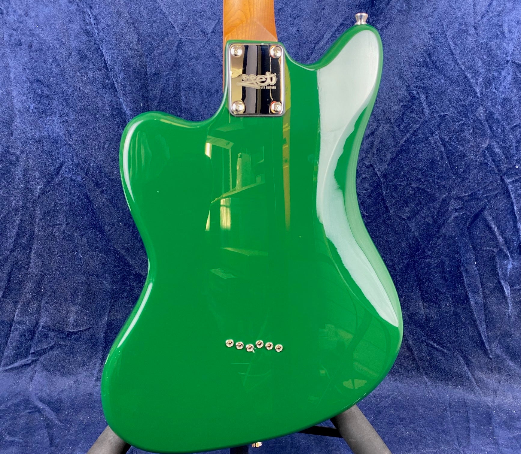 Green electric guitar body with a shiny finish, chrome neck plate, and six string ferrules on the back, set against a dark blue fabric background.