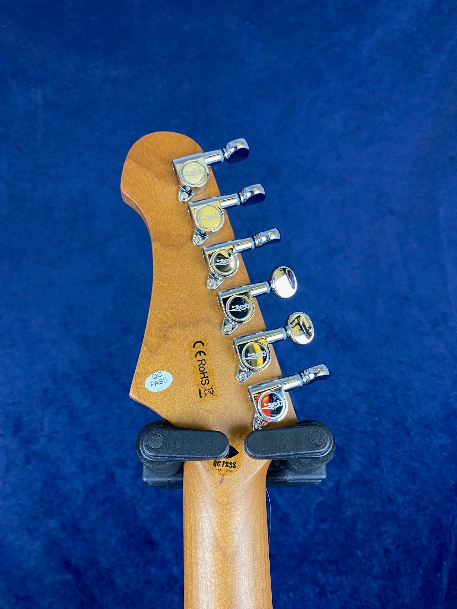 The back of an electric guitar headstock with six chrome tuning pegs, factory inspection stickers, and a wooden neck against a blue background.