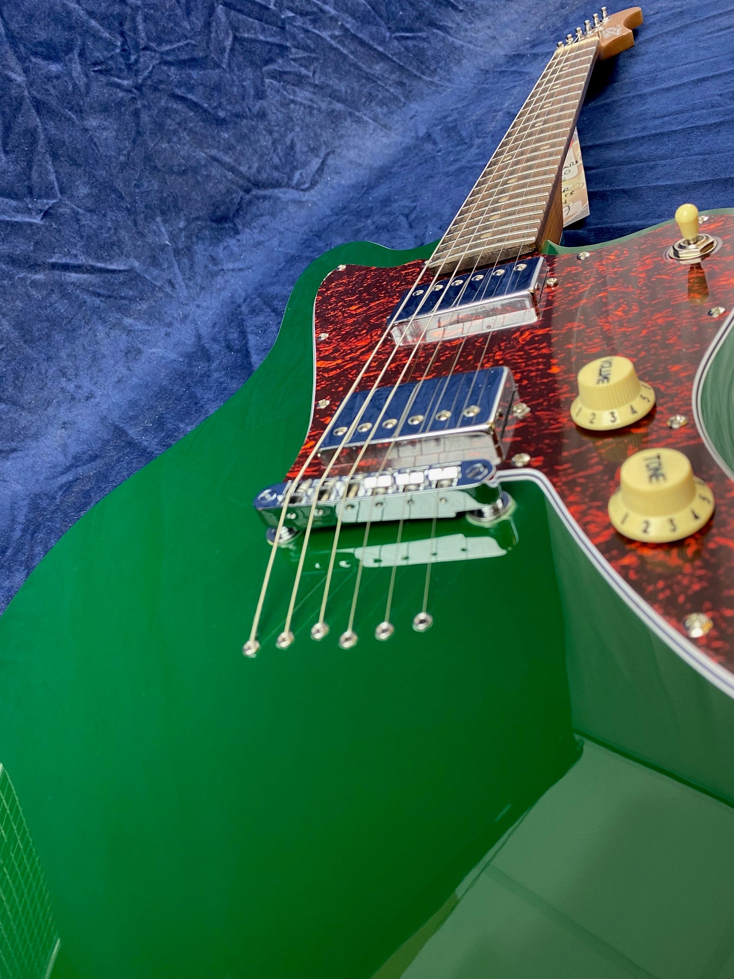 Green electric guitar with a red tortoiseshell pickguard, yellow control knobs labeled tone and volume, and metallic pickups against a blue background.