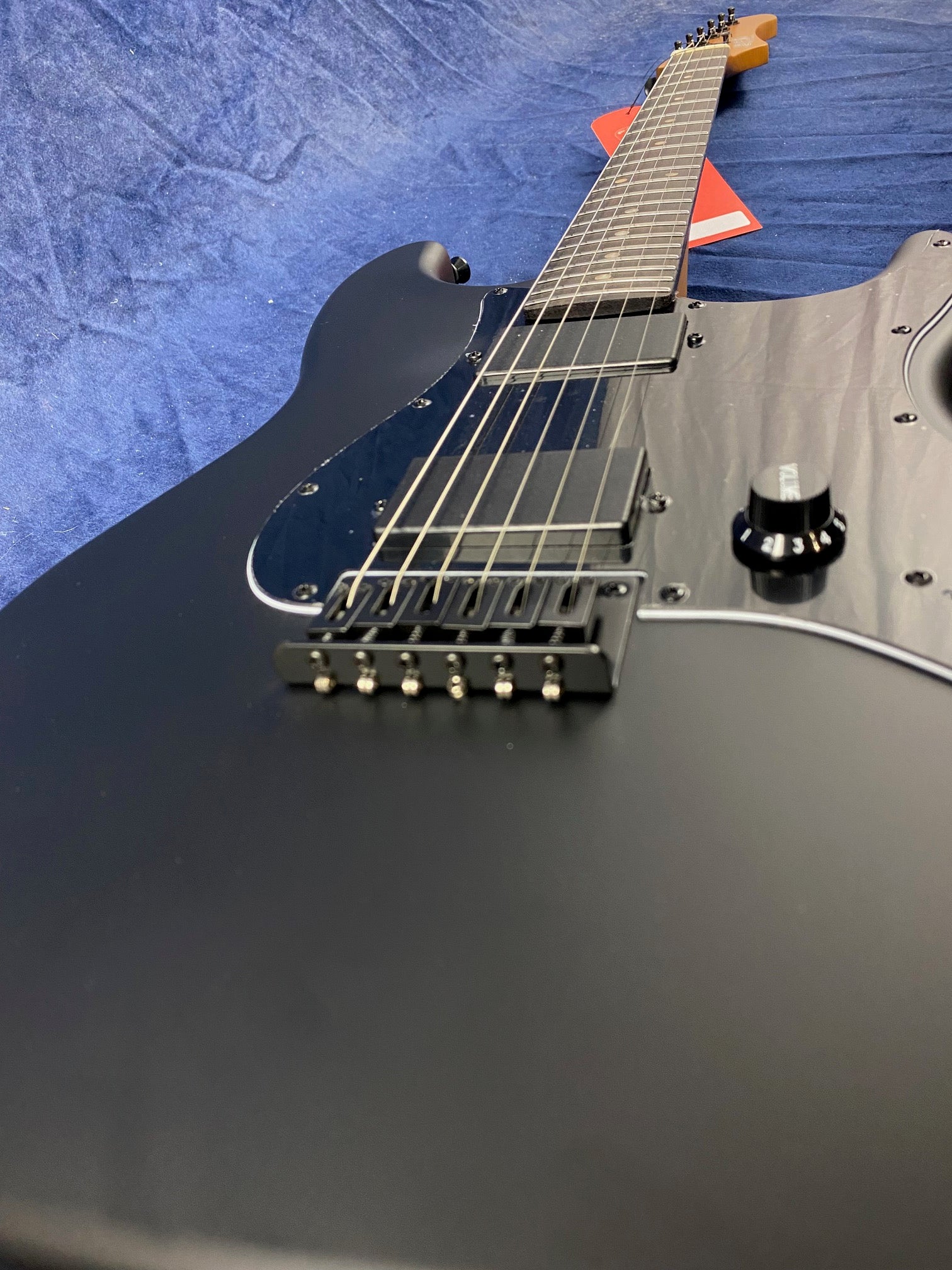 Matte black electric guitar with six strings, black pickups, a black pickguard, single volume knob, and a small red tag near the headstock.