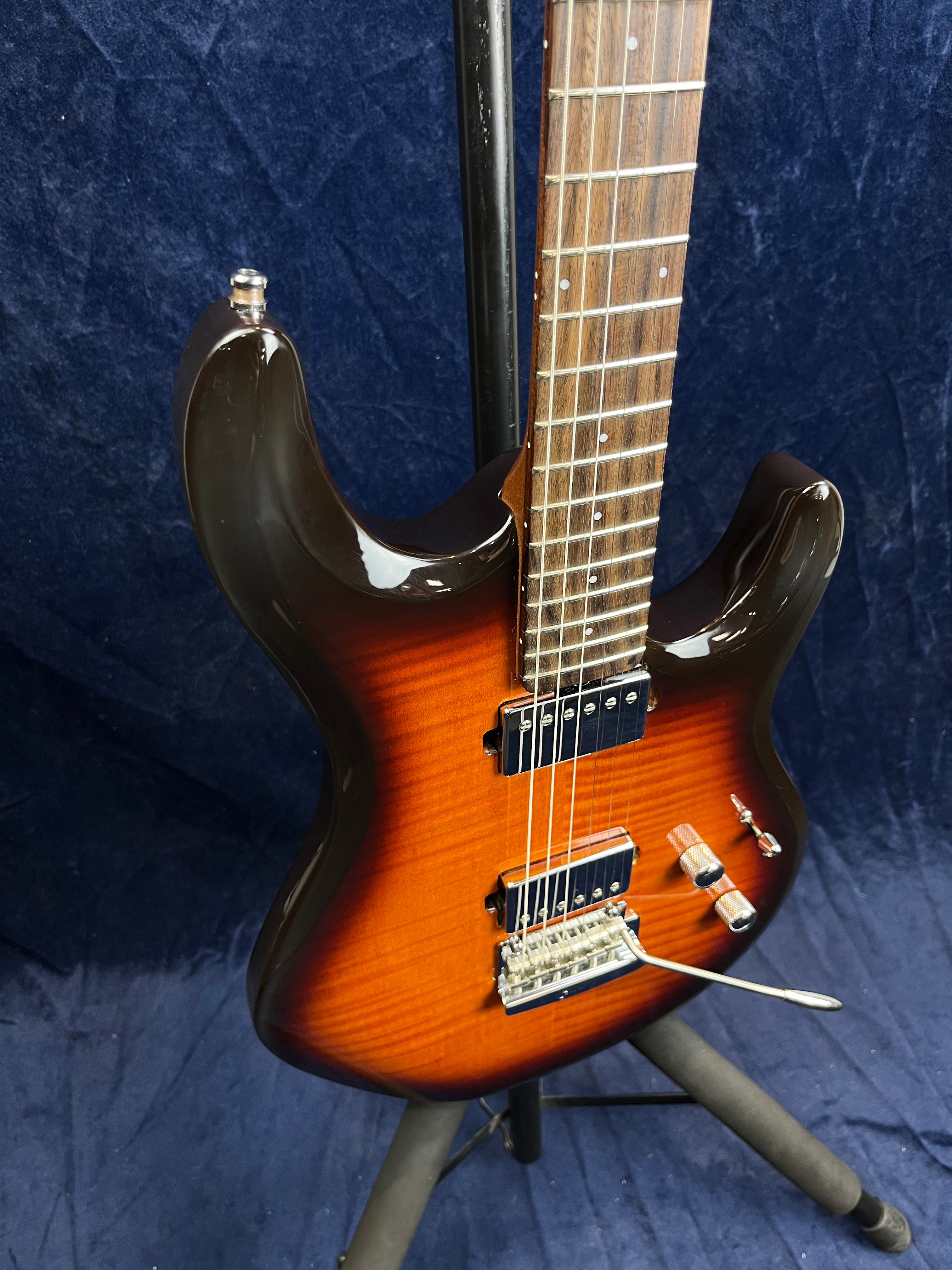 Guitare électrique à corps marron sunburst, deux micros doubles, vibrato, touches claires, posée sur un pied devant un fond bleu.