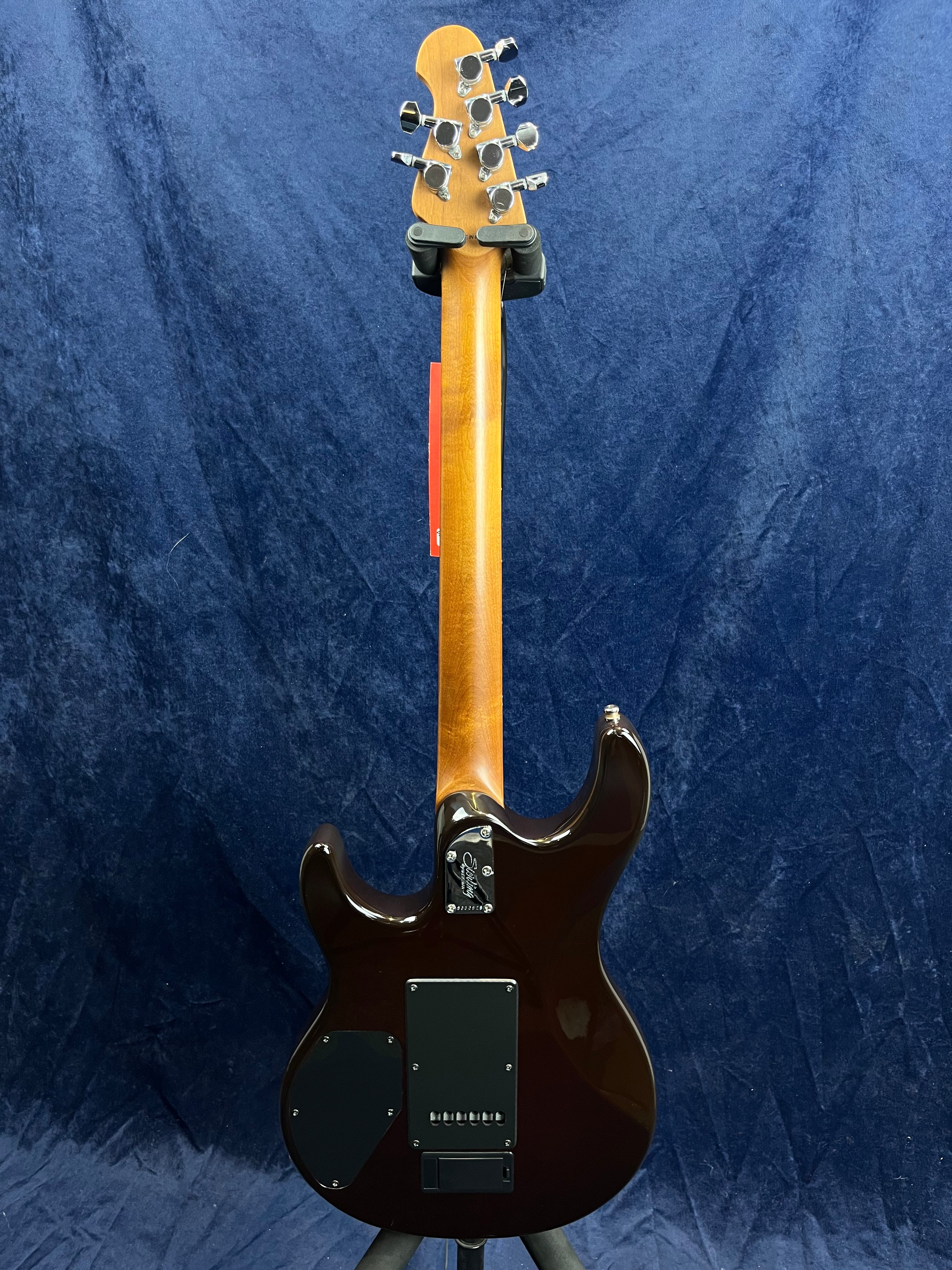 Back view of a dark brown electric guitar with a natural wood neck, chrome tuning pegs, and black electronics covers, standing against a blue backdrop.