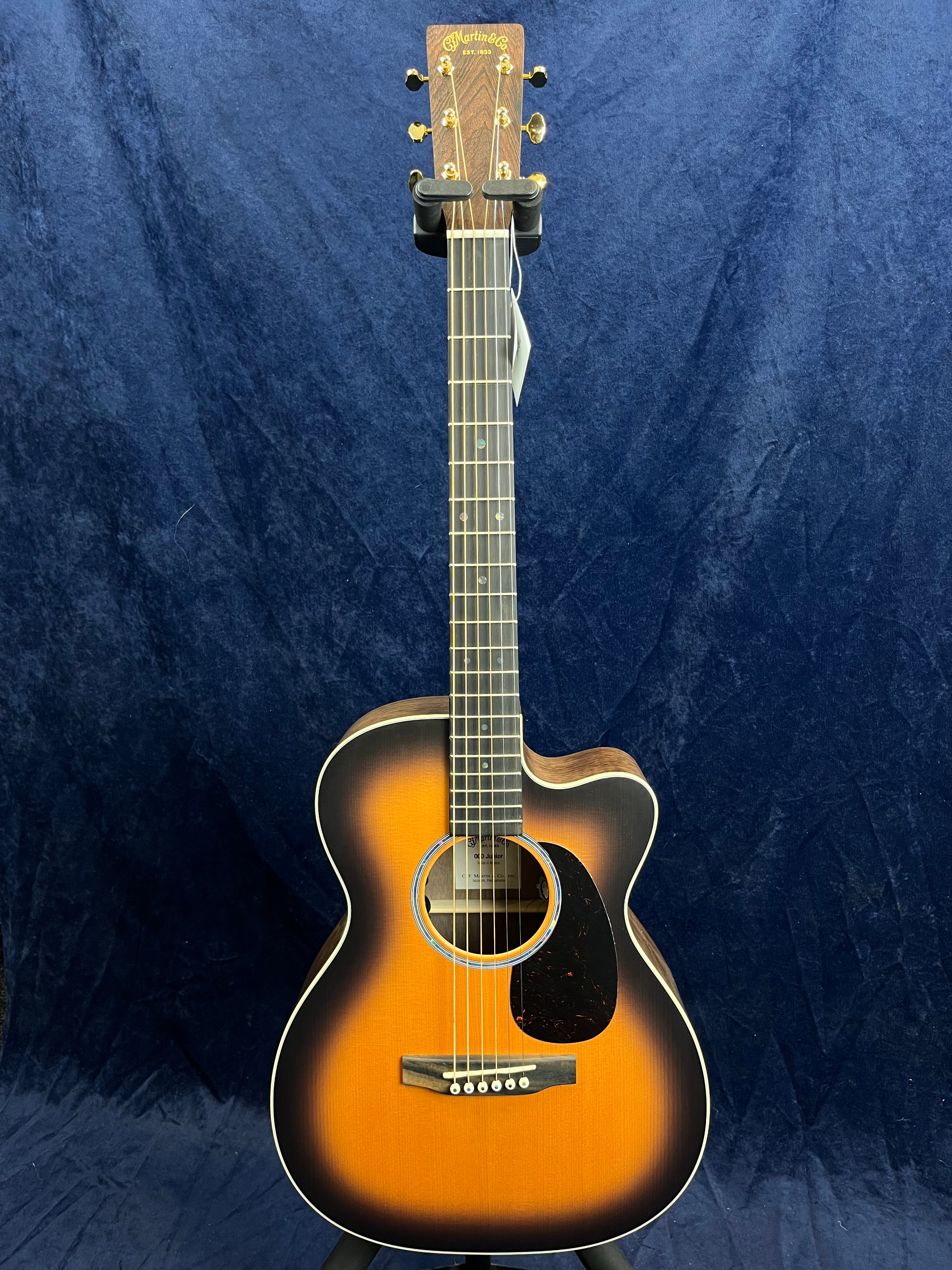 Martin acoustic guitar with sunburst finish, cutaway body, six strings, gold tuning pegs, and a black pickguard against a dark blue background.