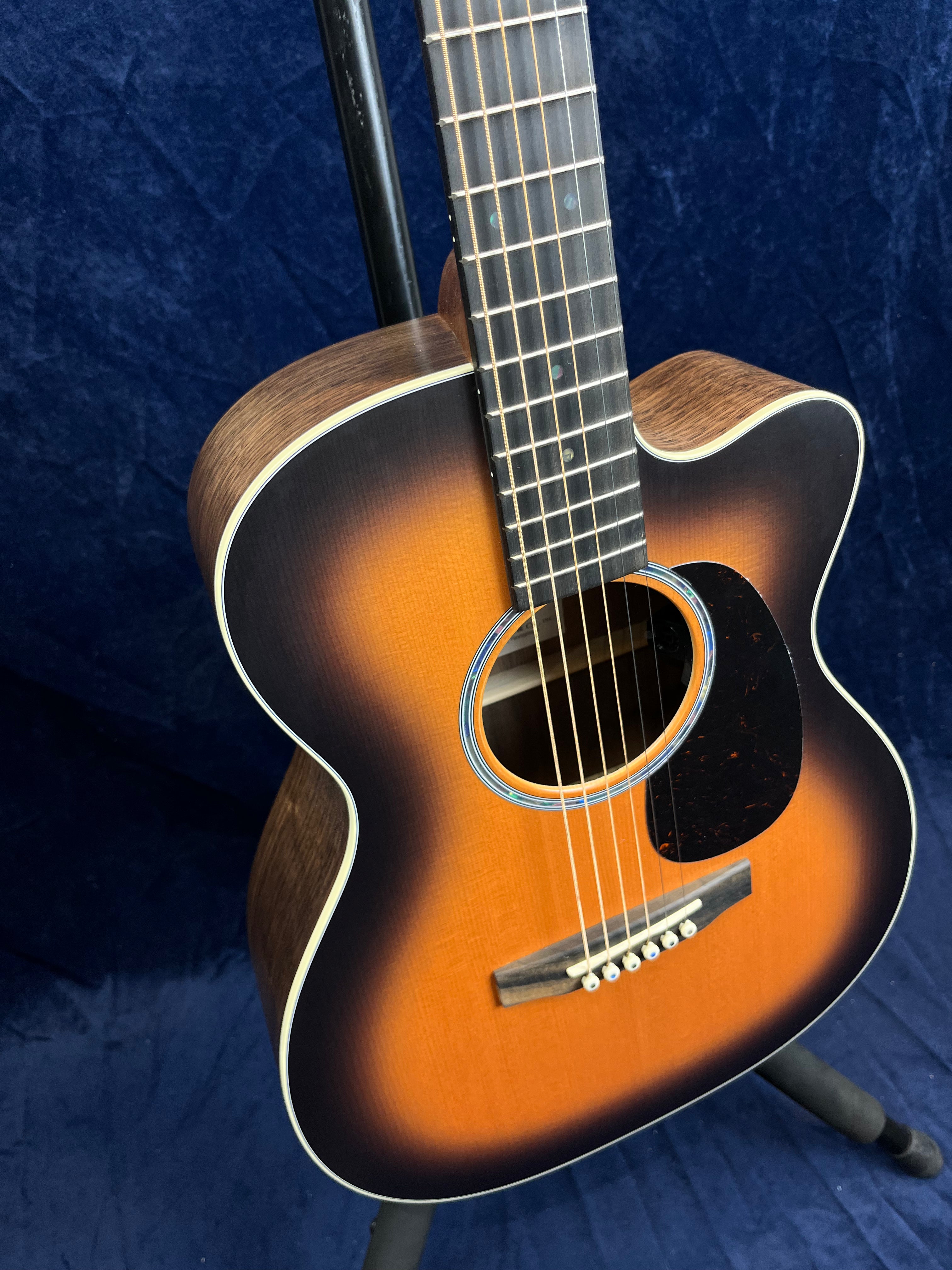 Martin acoustic guitar with a sunburst finish on the body, six strings, and a dark pickguard, resting on a stand against a dark blue background.