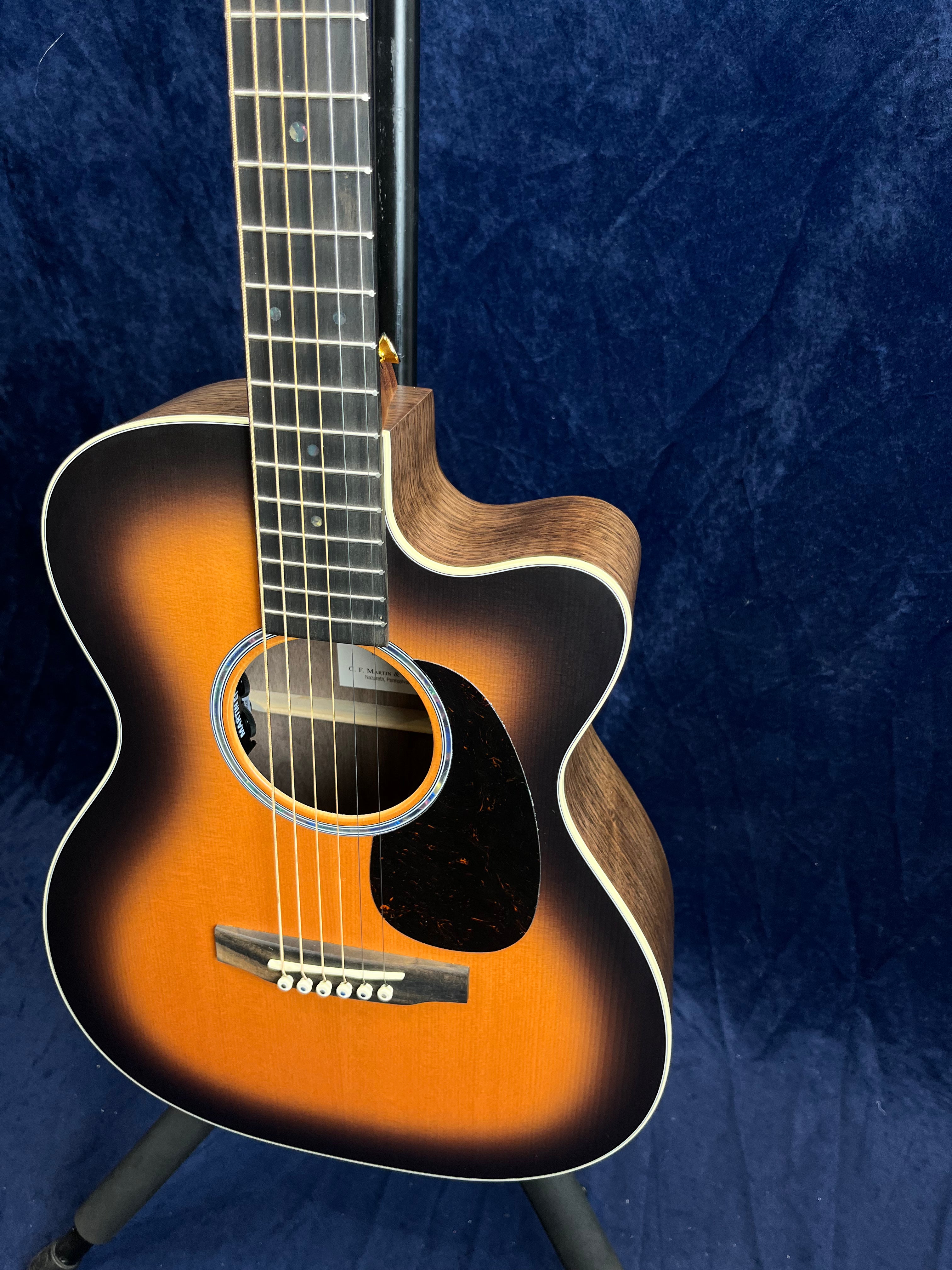 Martin acoustic guitar with a sunburst finish, cutaway body, white binding, tortoiseshell pickguard, and six strings, displayed on a stand.