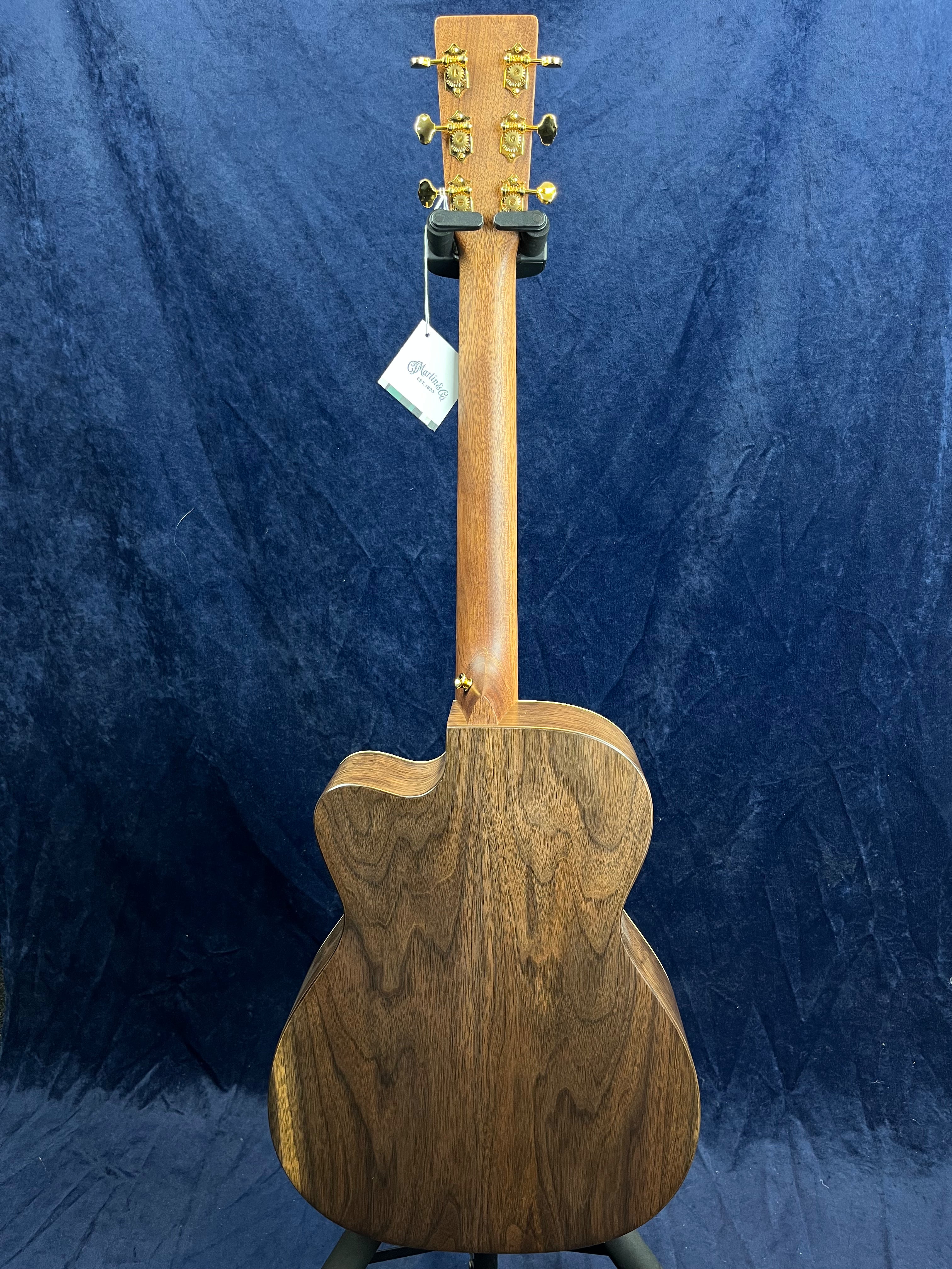 Acoustic guitar viewed from the back showing dark wood grain, gold tuning pegs, and a hanging tag attached to the headstock.