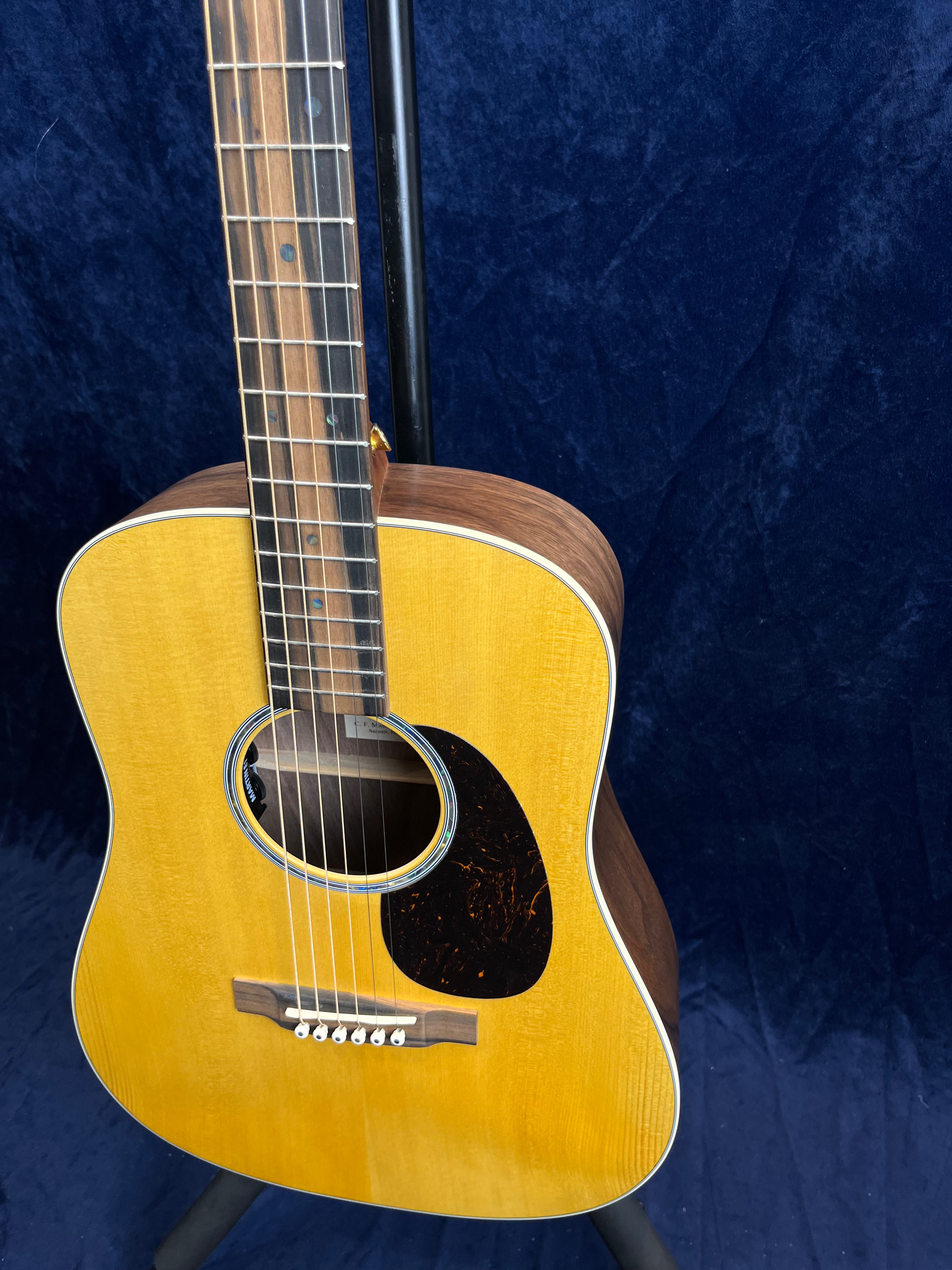 Acoustic guitar with a light wood top, dark pickguard, and six strings, displayed on a stand against a dark blue background.