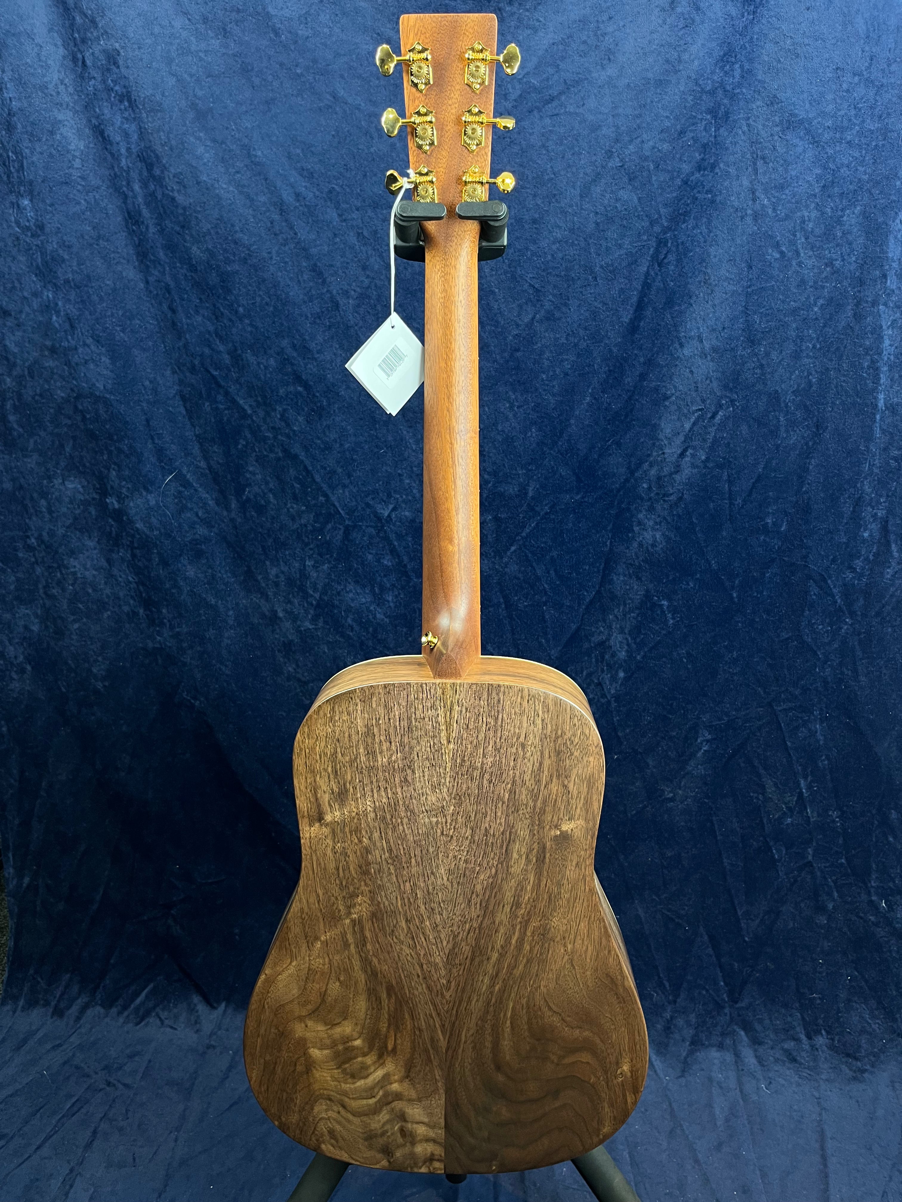 Acoustic guitar shown from the back, displaying a rich, dark wood grain body, gold tuners, and a tag hanging from the headstock against a dark background.