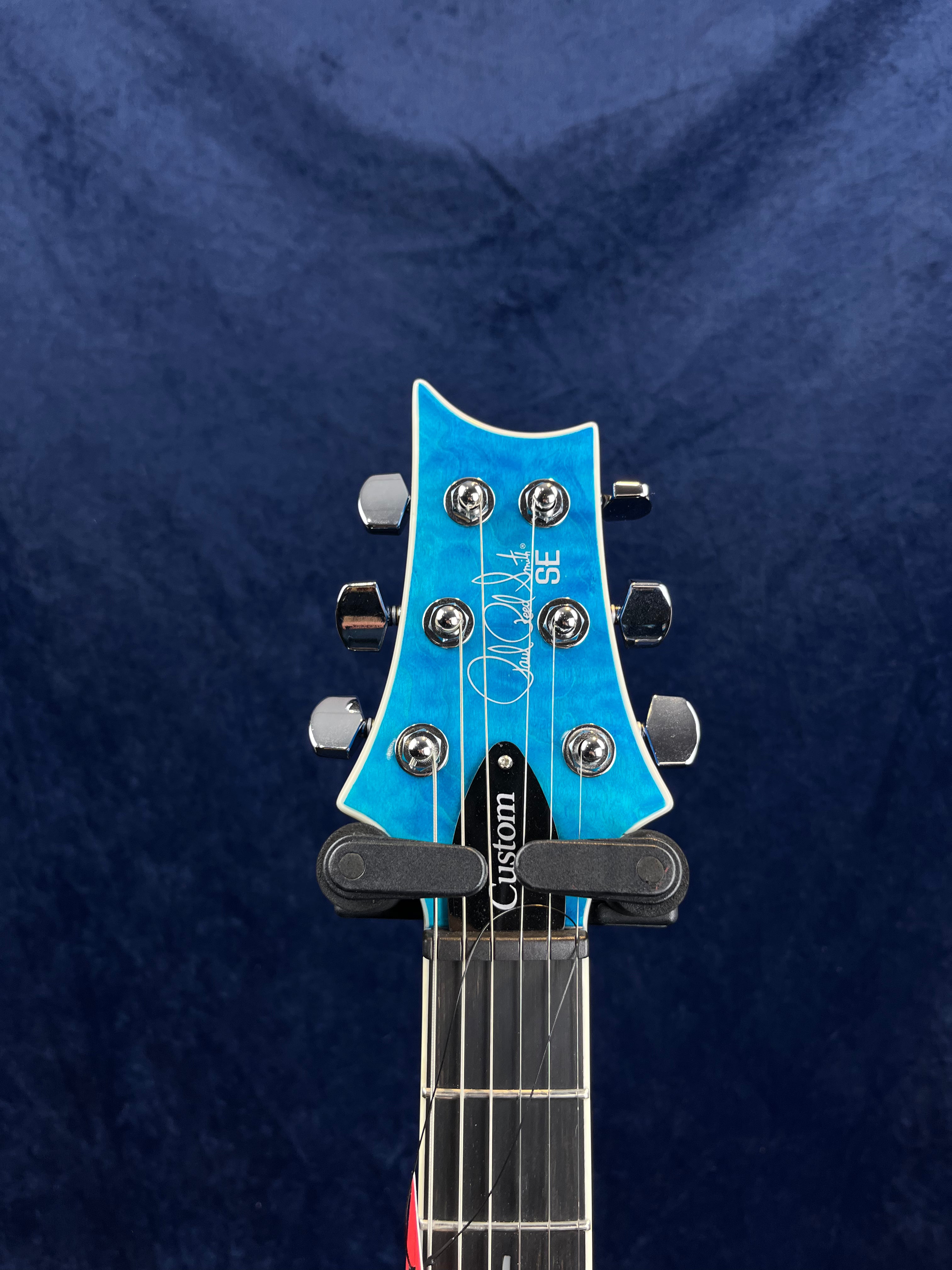 Blue electric guitar headstock with six chrome tuning pegs, "SE" and "Custom" markings, and a signature-style logo on the front.