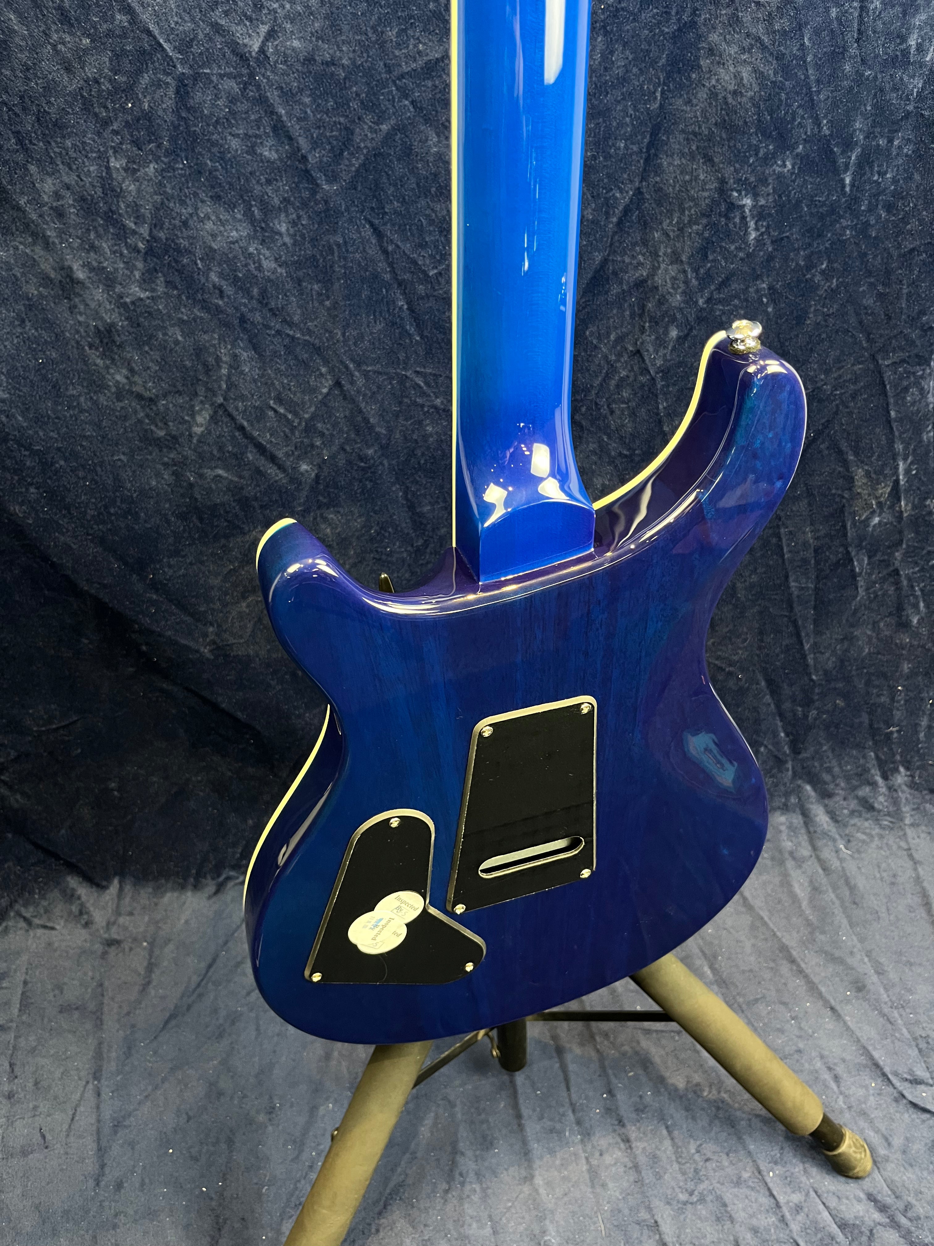 Electric guitar with a glossy blue finish shown from the back, featuring two black plastic cavity covers, standing on a tripod guitar stand.