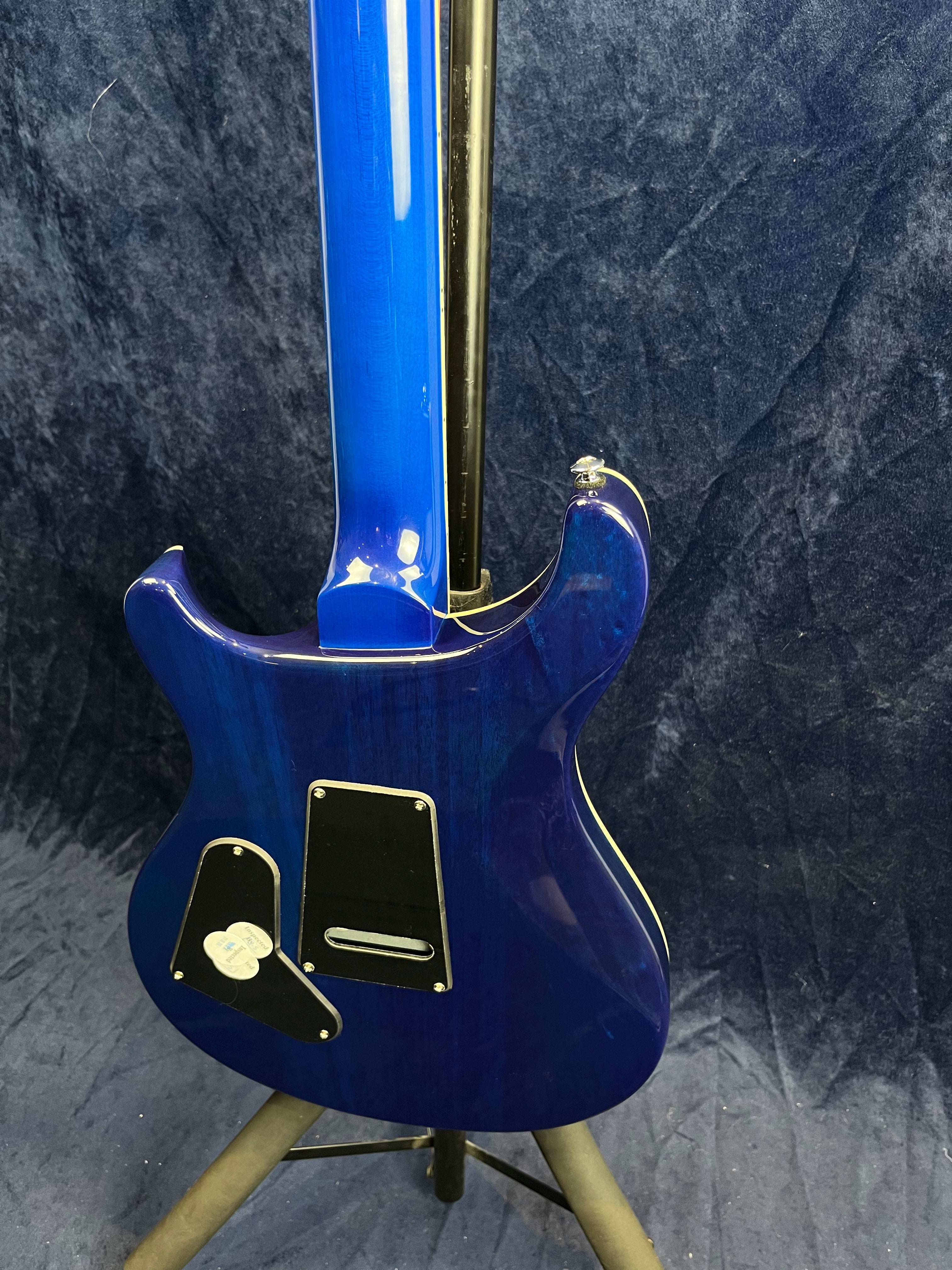 Bright blue electric guitar shown from the back, with black plastic cavity covers and a guitar stand, set against a dark textured background.