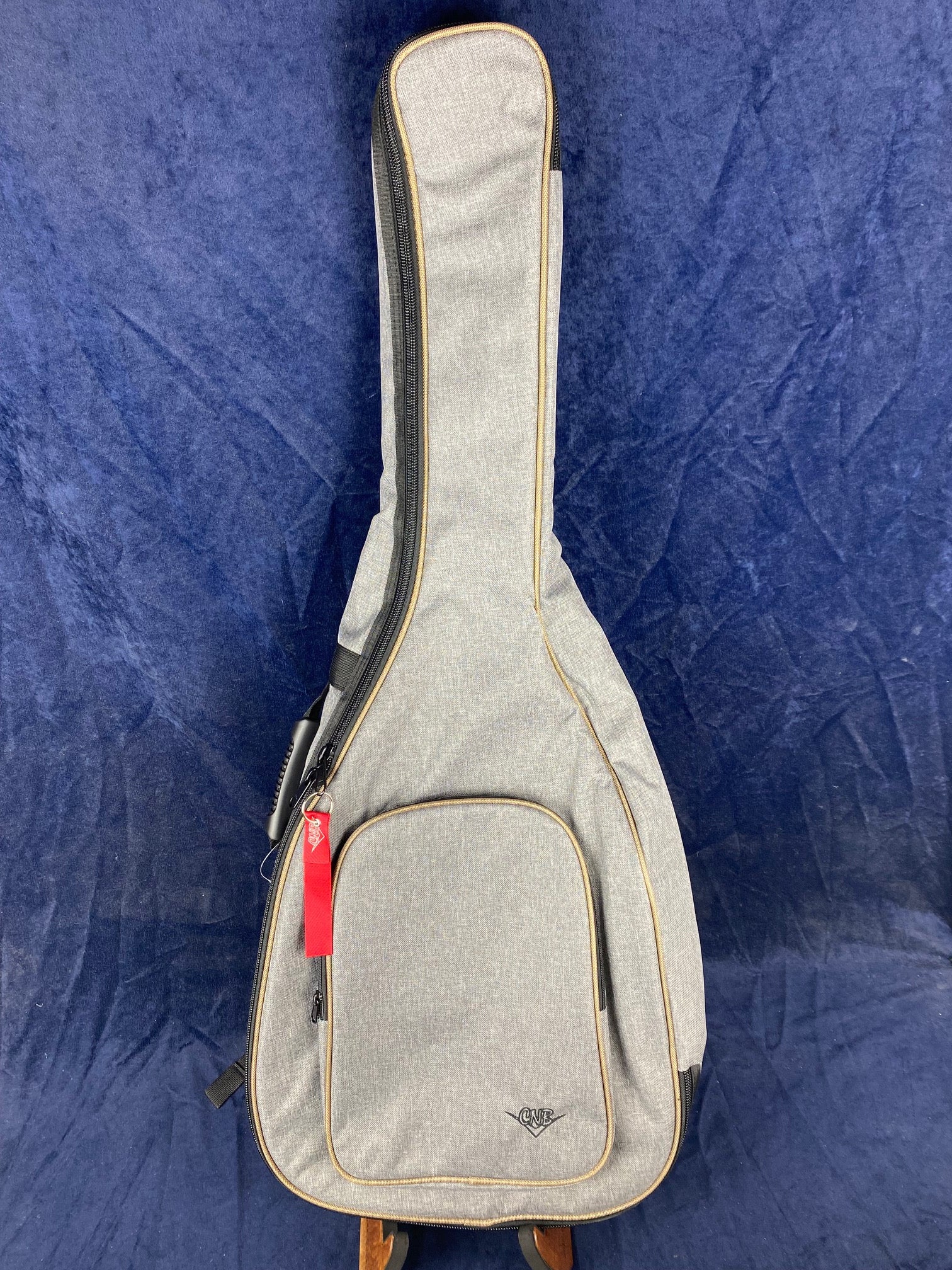 A light gray soft acoustic guitar case with tan trim, front zippered pocket, and a red keychain tag, standing upright against a dark blue background.