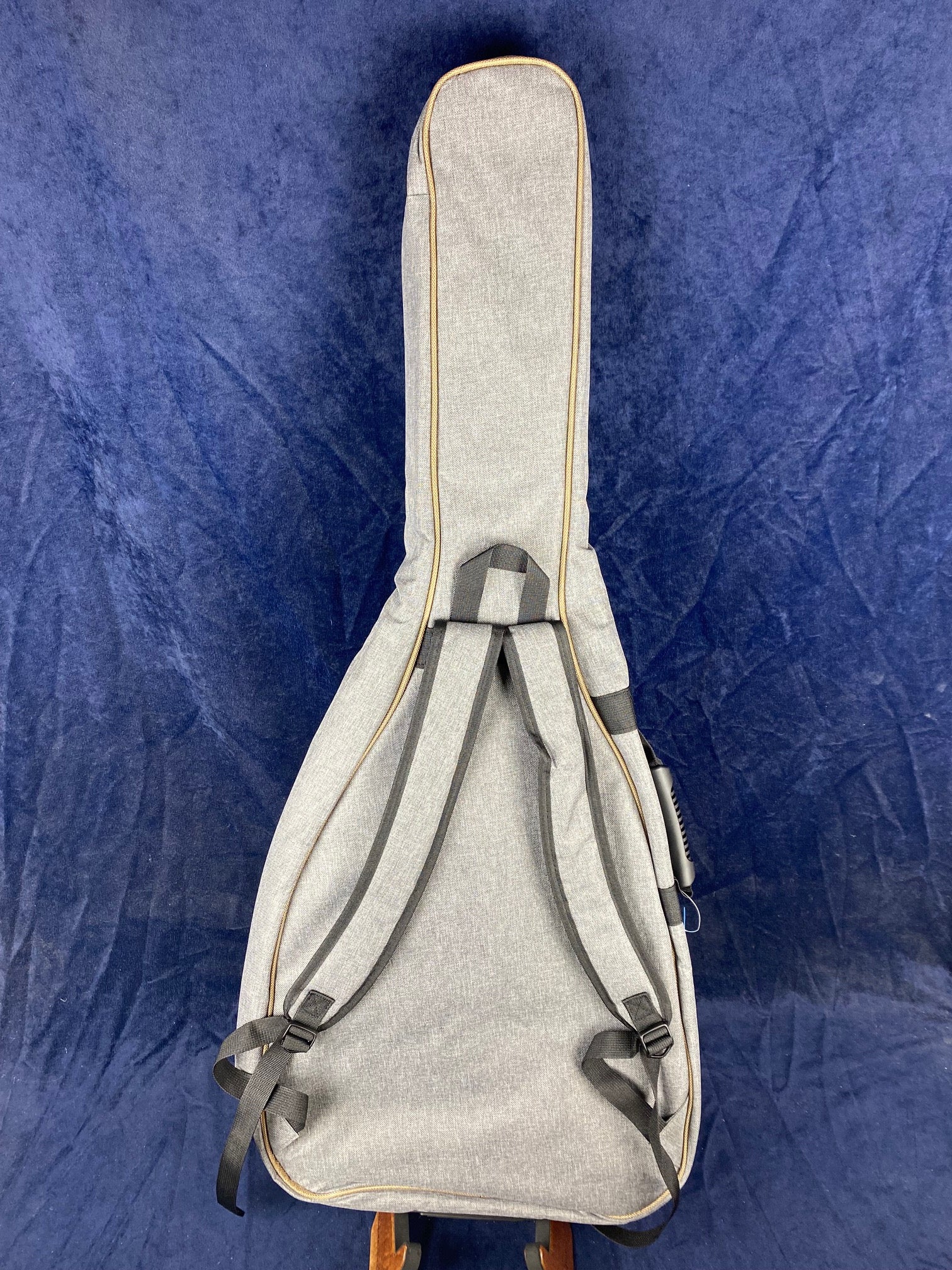 Light gray soft case for an acoustic guitar, shown upright with black adjustable backpack straps against a dark blue background.