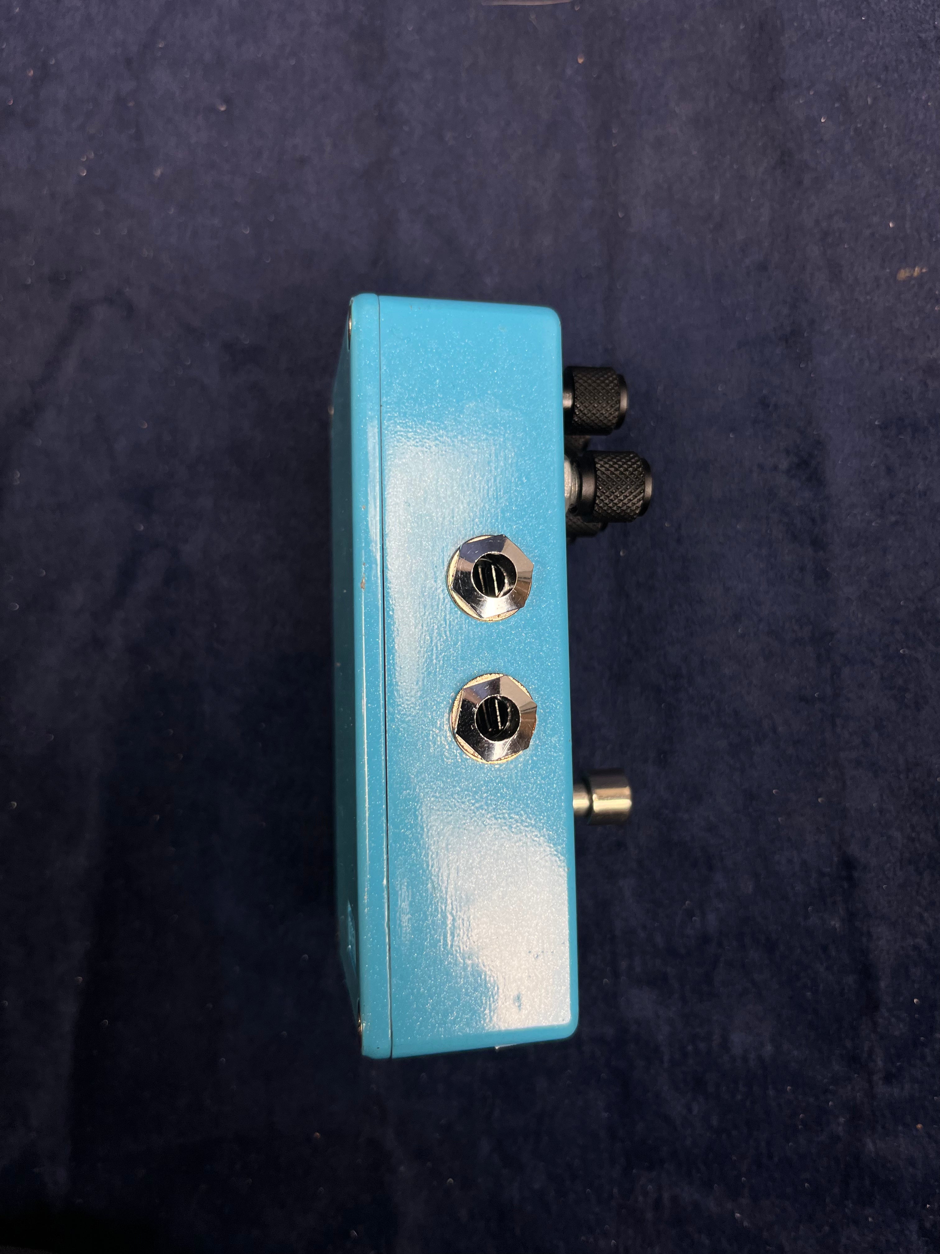 Light blue FX pedal with two metal audio jacks on the side, two black control knobs, and a power jack, shown against a dark background.