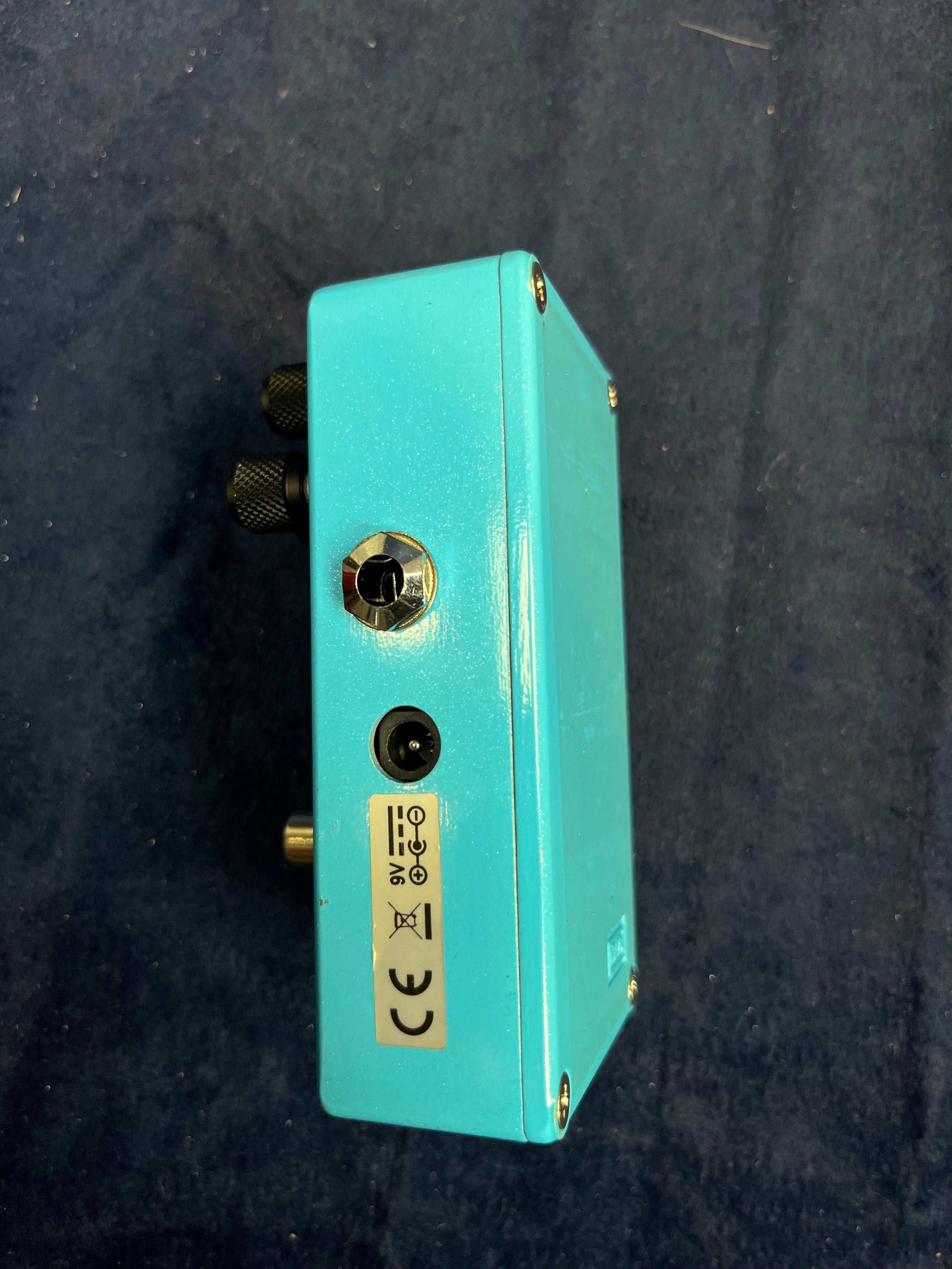 Light blue FX pedal showing its side, featuring a 9V power input, jack input, and a silver CE certification sticker on a dark background.