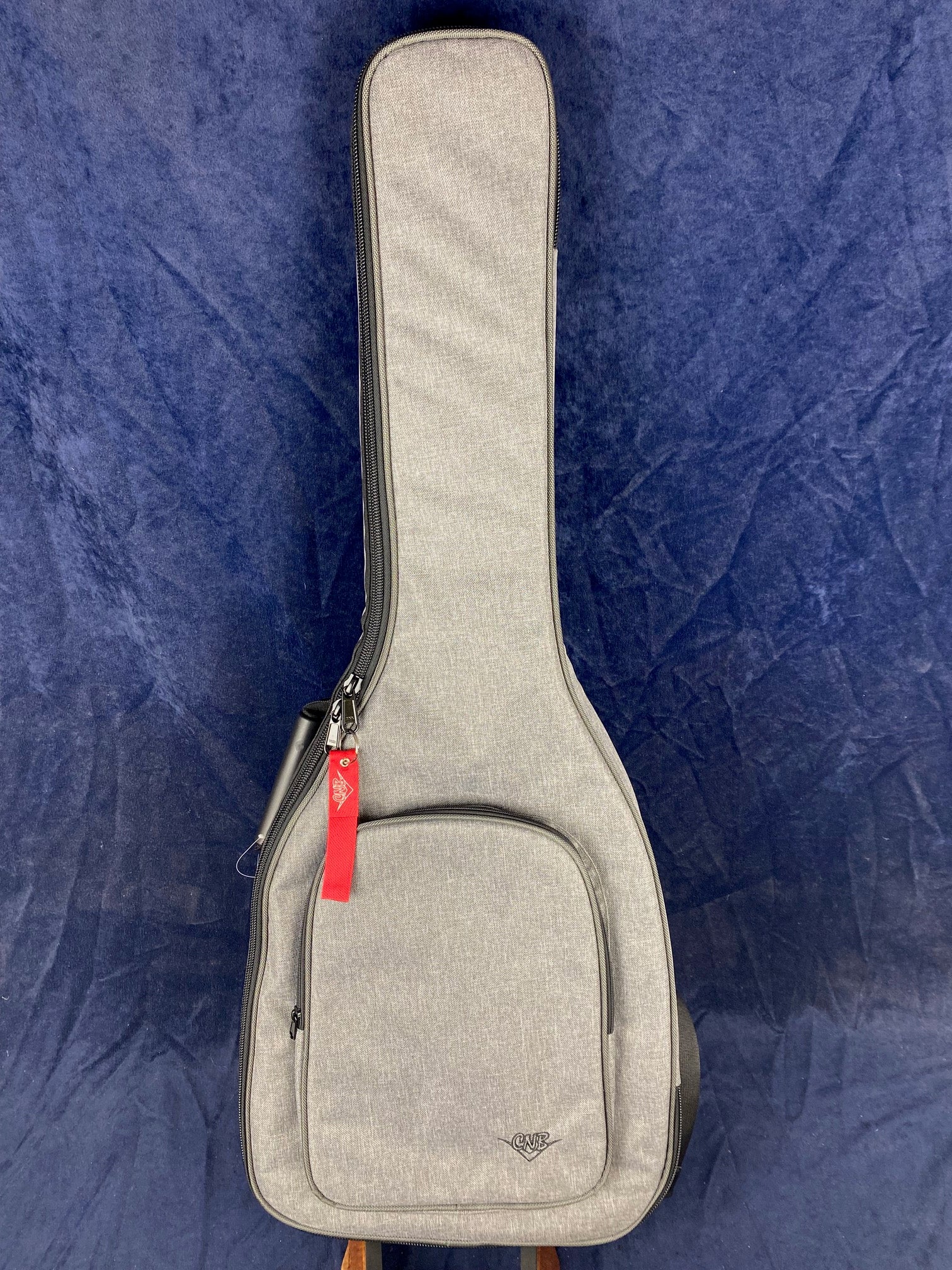 A light gray electric guitar gig bag with a front zippered pocket, a black zipper, and a red tag attached near the top.