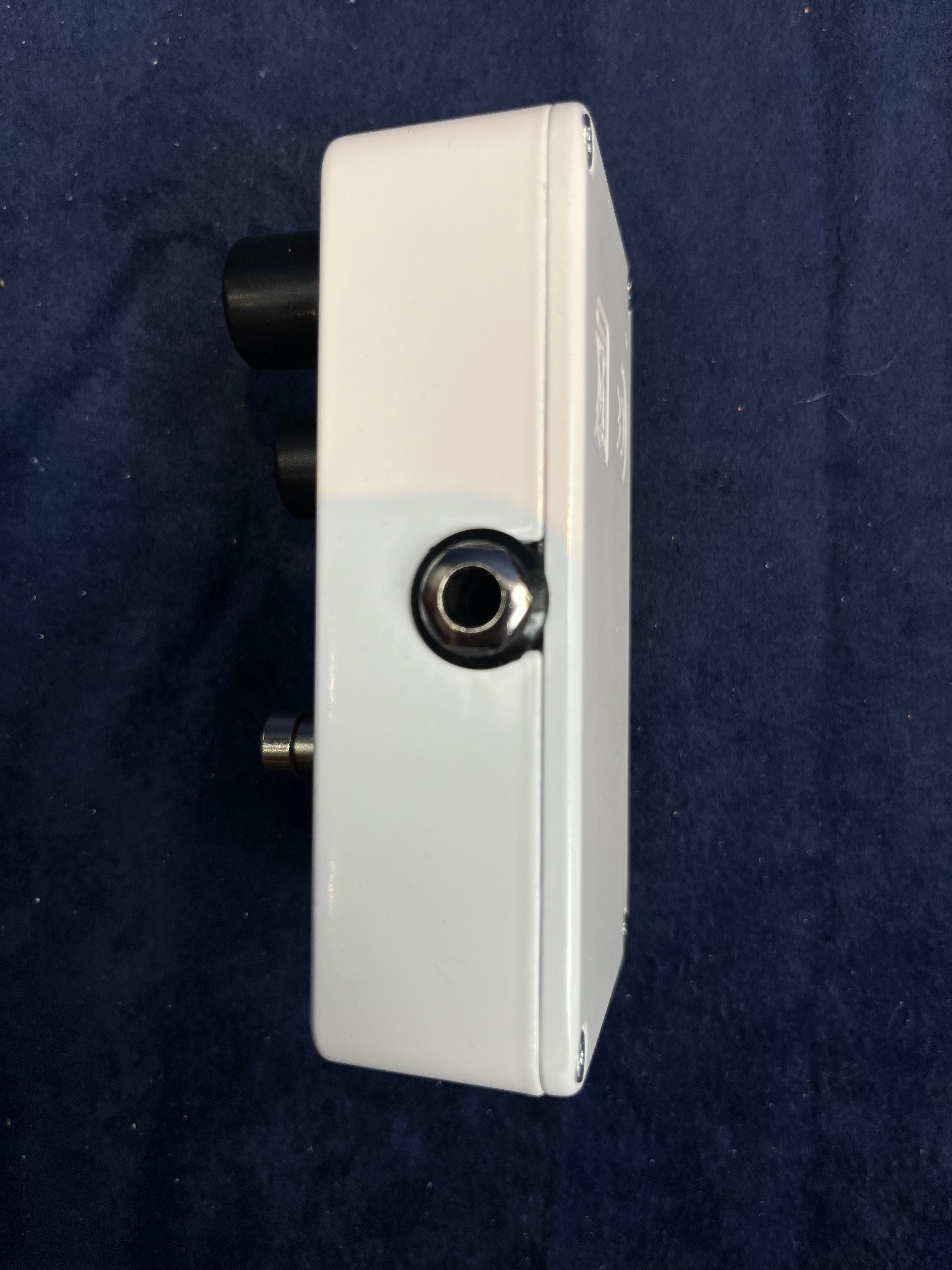 Boîtier d’effet pour guitare, blanc, vu de côté avec une prise jack métallique et deux gros boutons noirs visibles à l’arrière.