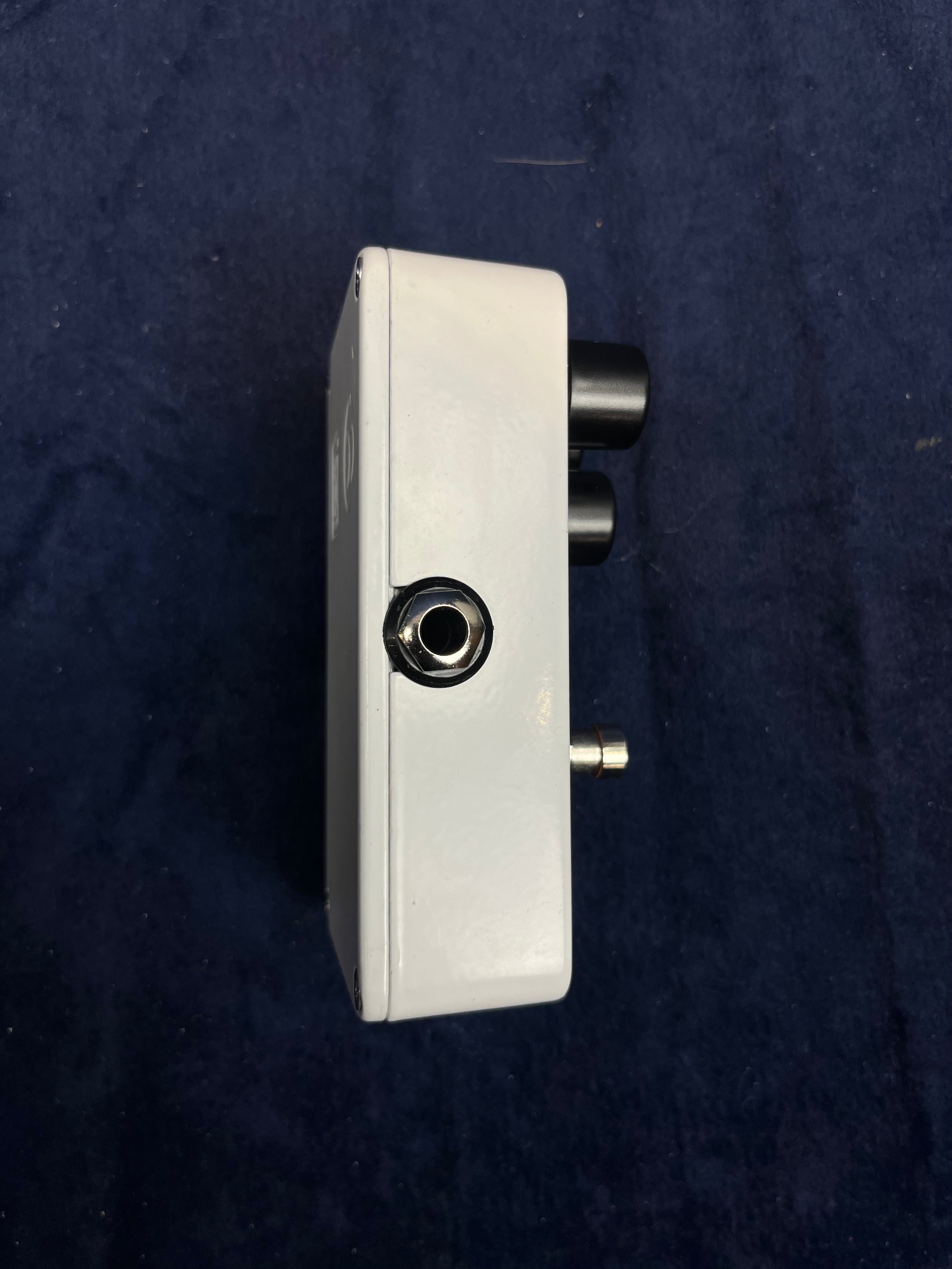 White FX pedal shown sideways; features one jack input, two black control knobs, and a small metal switch on a dark fabric background.