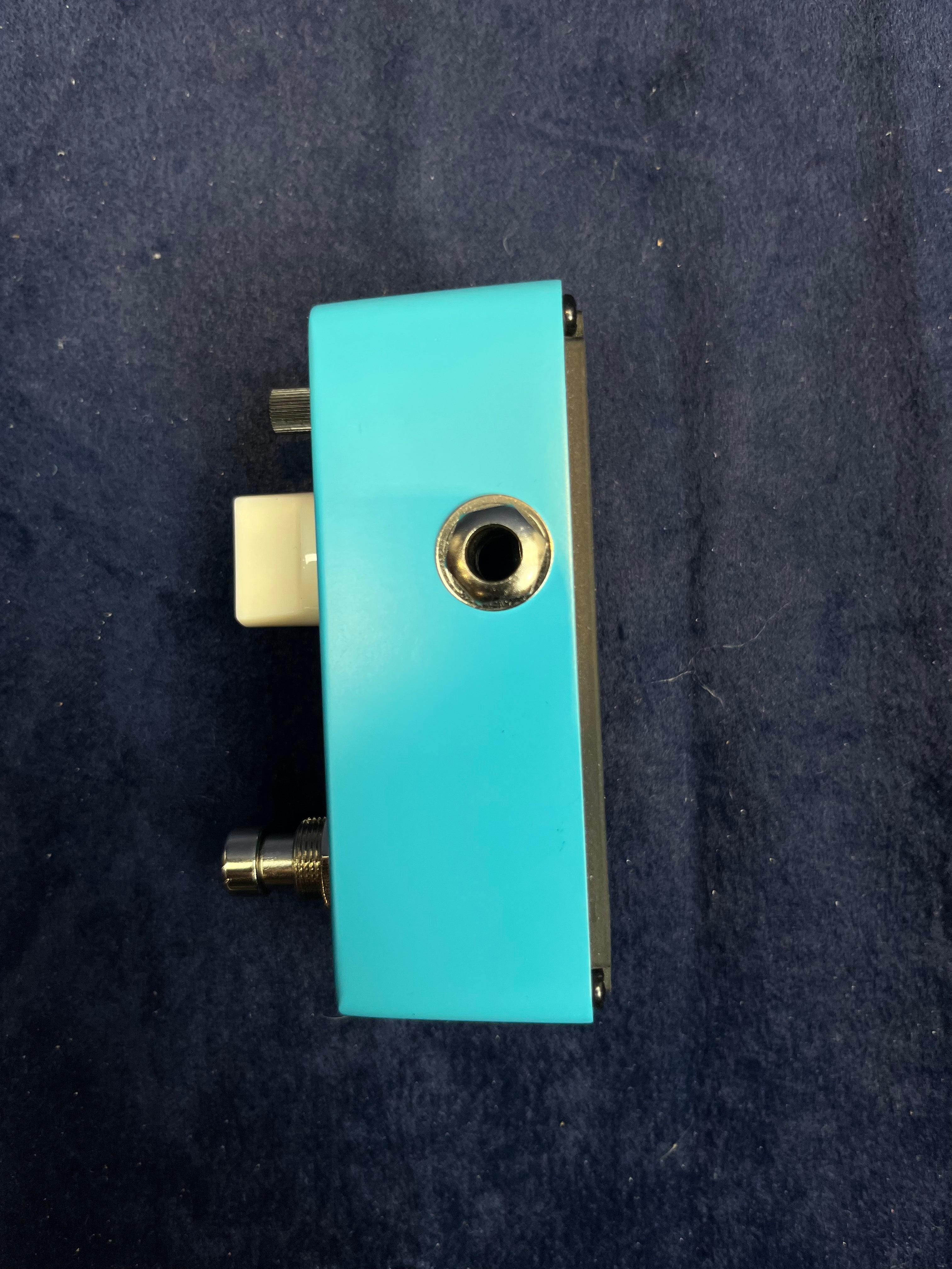 Compact FX pedal in bright blue with a single metal input jack, a footswitch, and a small knob, displayed on a dark fabric surface.