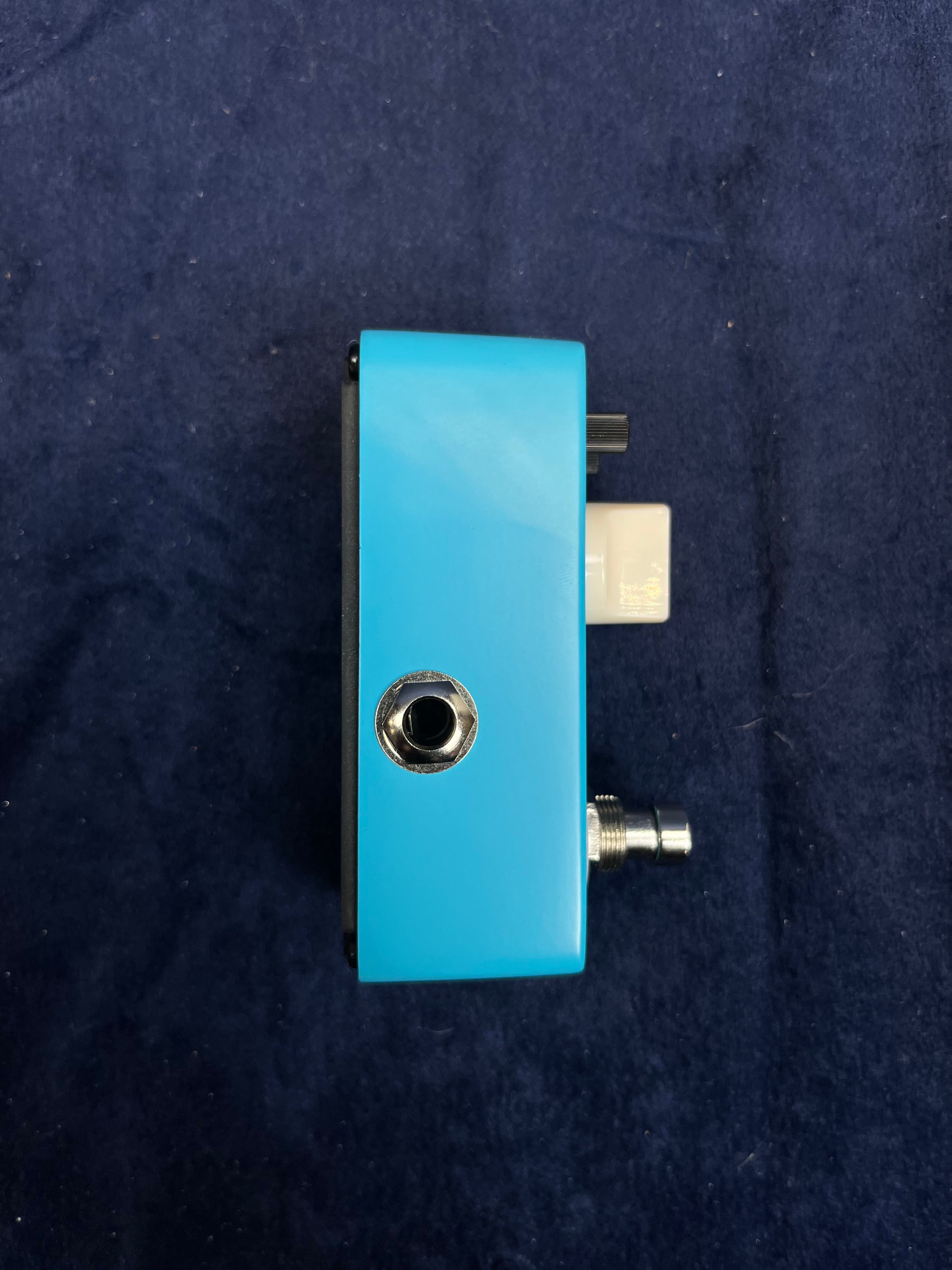 A compact blue FX pedal seen from the side, showing a single input jack, a white knob, and a metal footswitch against a dark blue background.