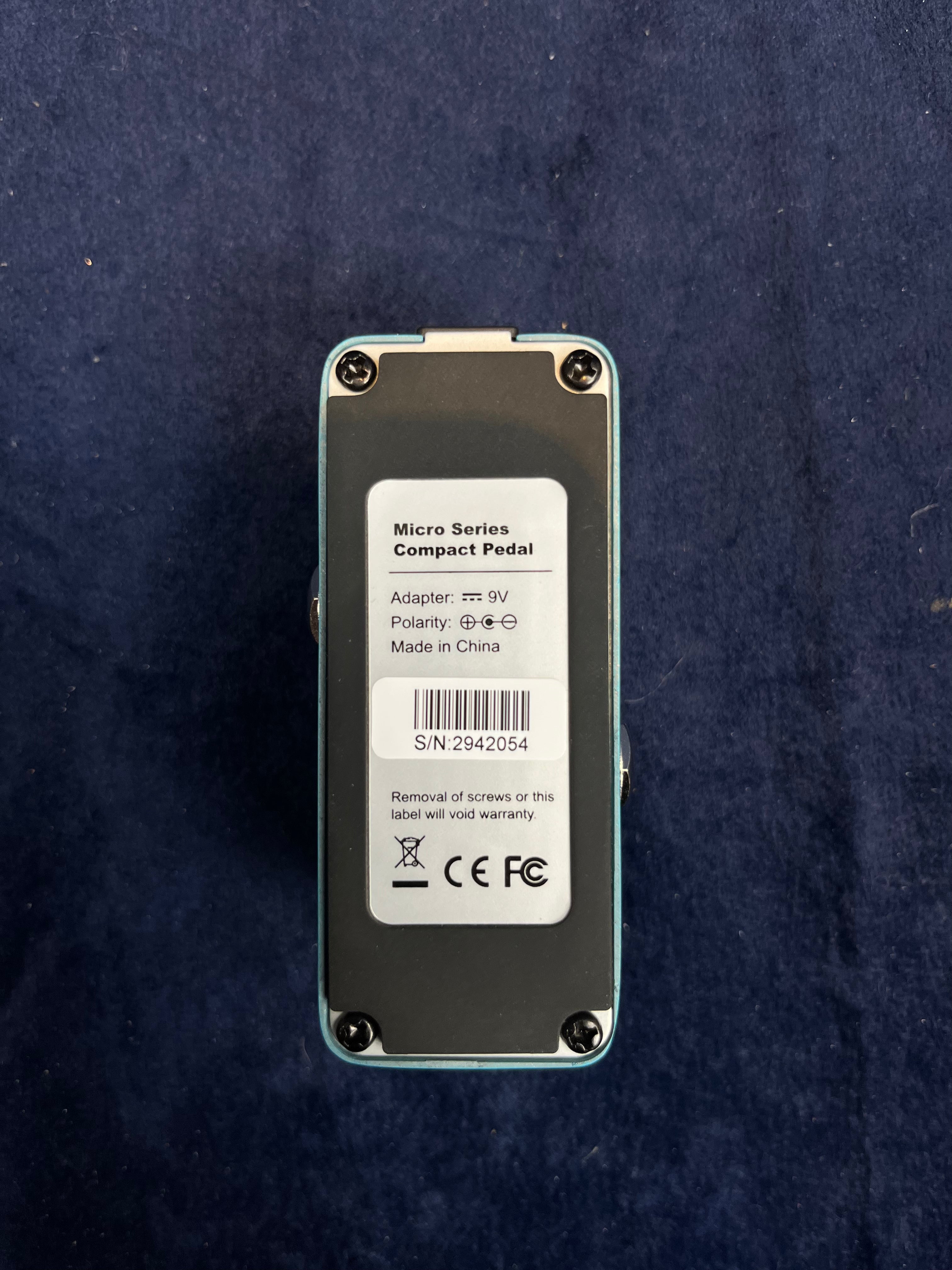 Pédale d’effet compacte rectangulaire, étiquette blanche précisant alimentation 9V, numéro de série et certification CE et FCC au dos.