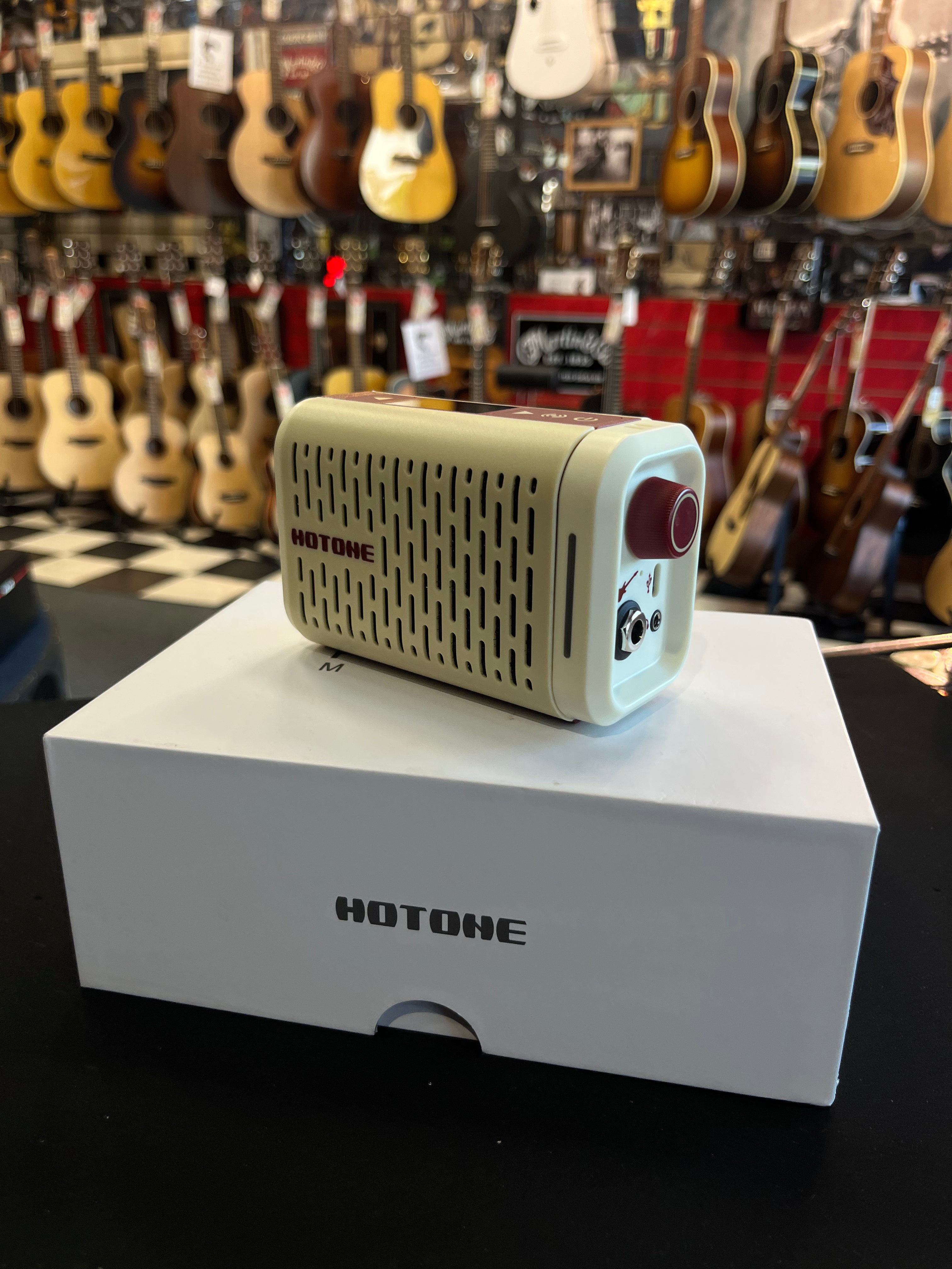 Compact cream and red Hotone amplifier with vented grill, headphone jack, and control knob, sitting on a branded white box in a guitar shop.