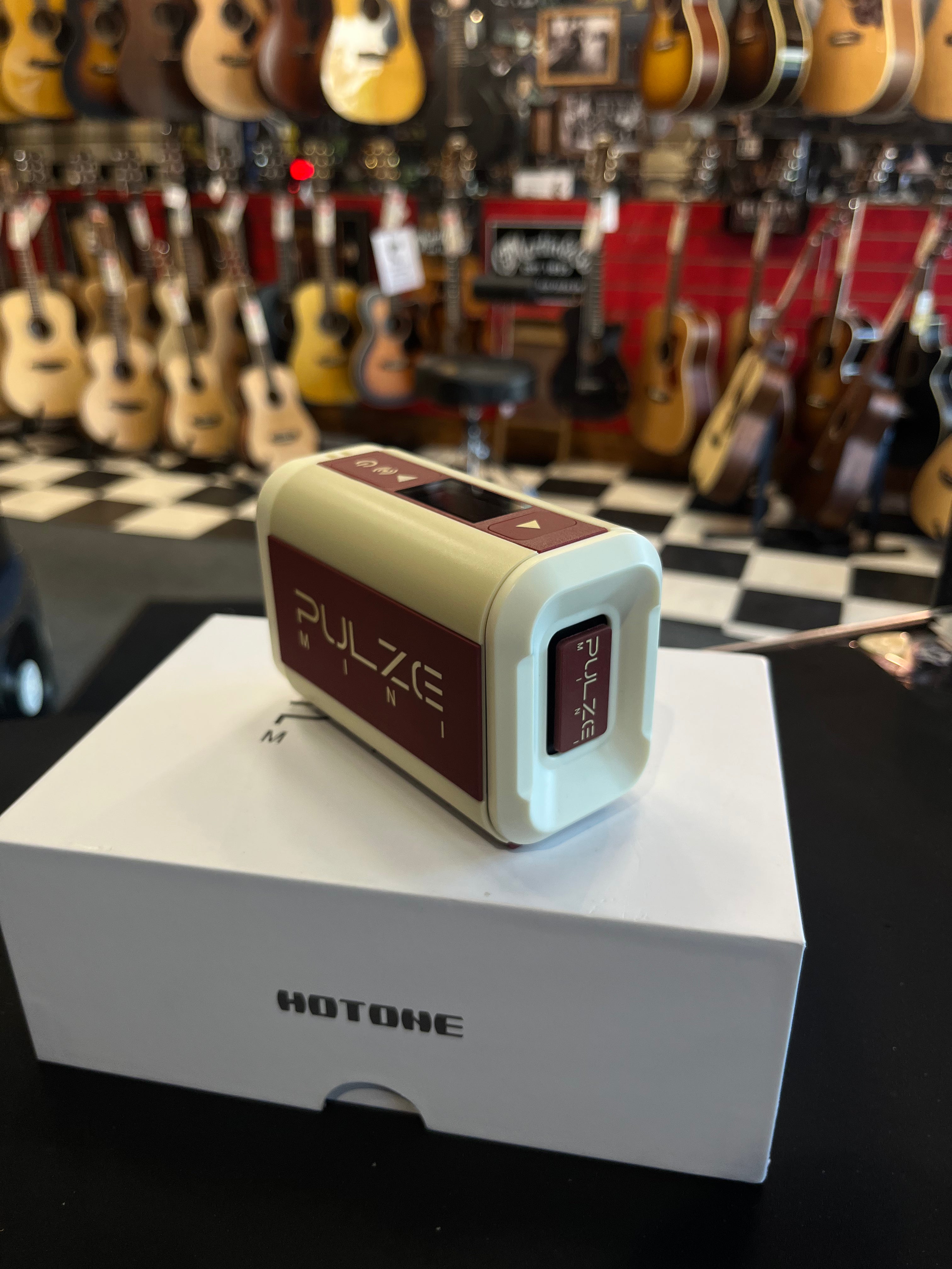 Compact Hotone Pulse Mini amplifier, cream and maroon, sits on its box in a guitar store with many acoustic guitars hanging in the background.