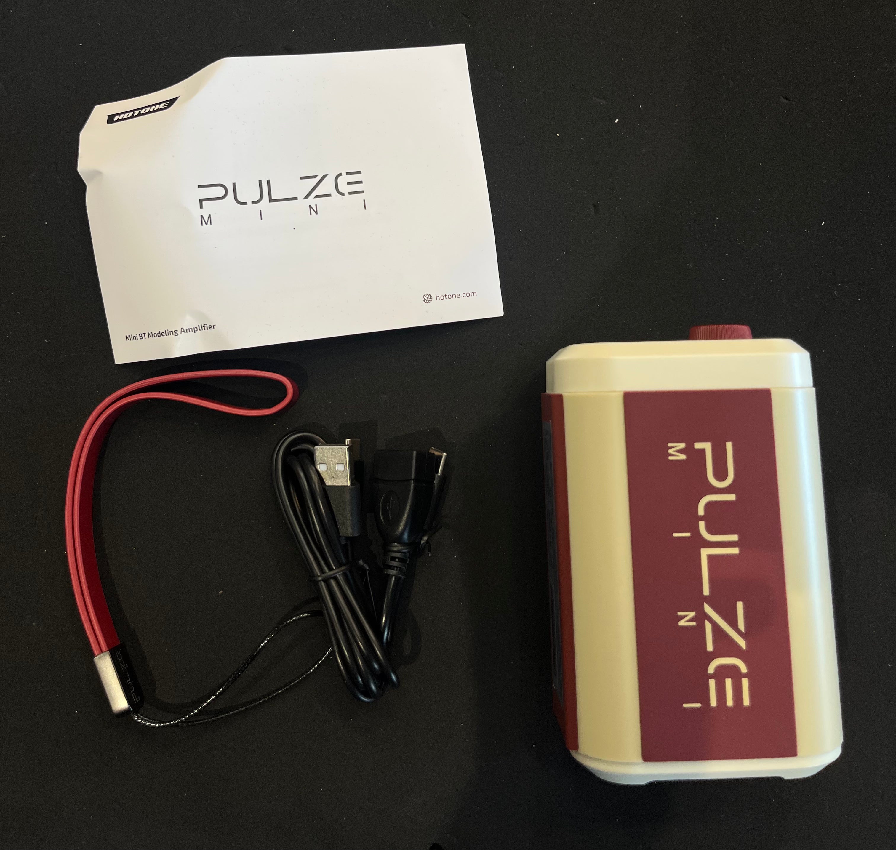 PULZE Mini amplifier, rectangular with red and cream colors, shown with a manual, USB cable, and red wrist strap on a black surface.