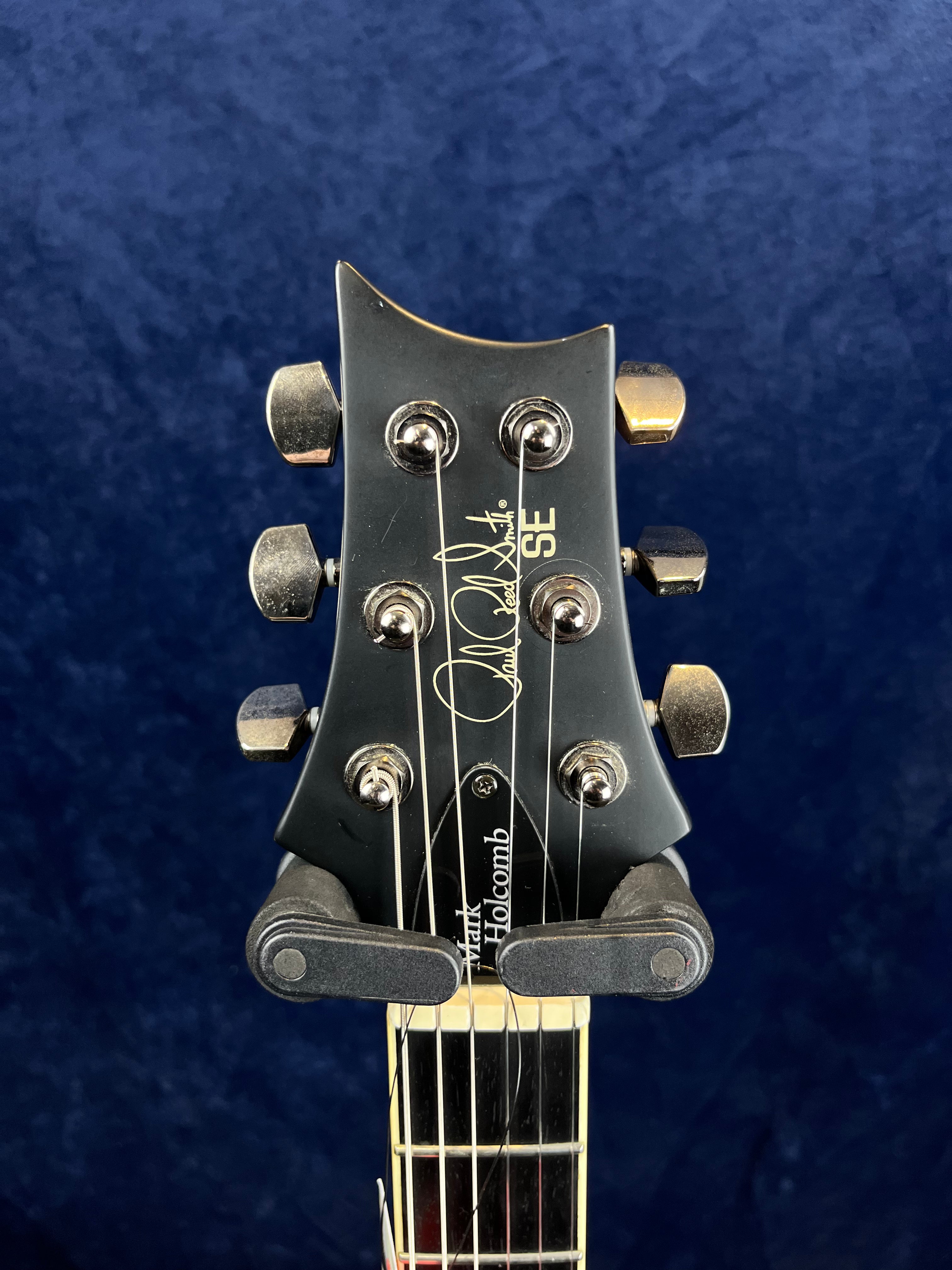 Black electric guitar headstock with six tuning pegs, PRS SE logo, Paul Reed Smith signature, and "Mark Holcomb" written near the truss rod cover.