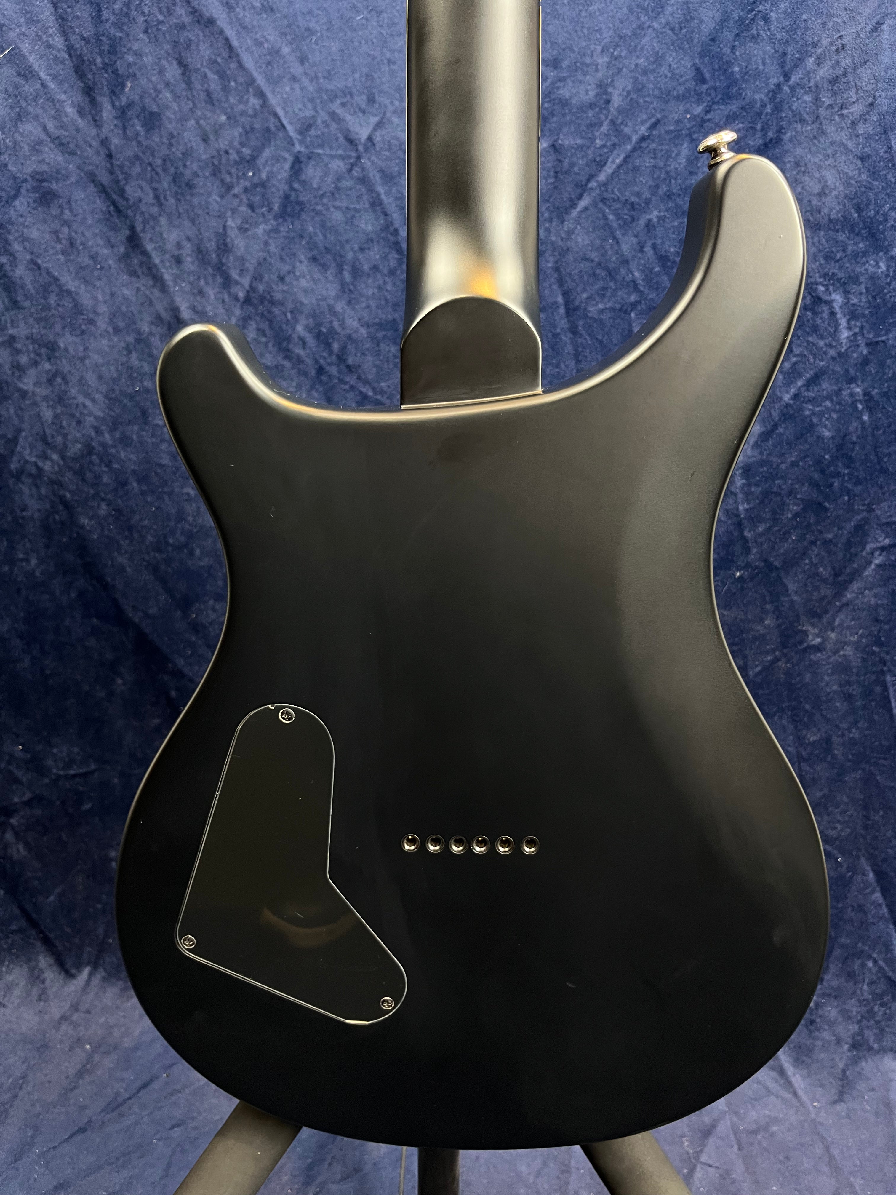Matte black electric guitar photographed from the back; features a curved body, rear control cavity cover, and string ferrules in a line.